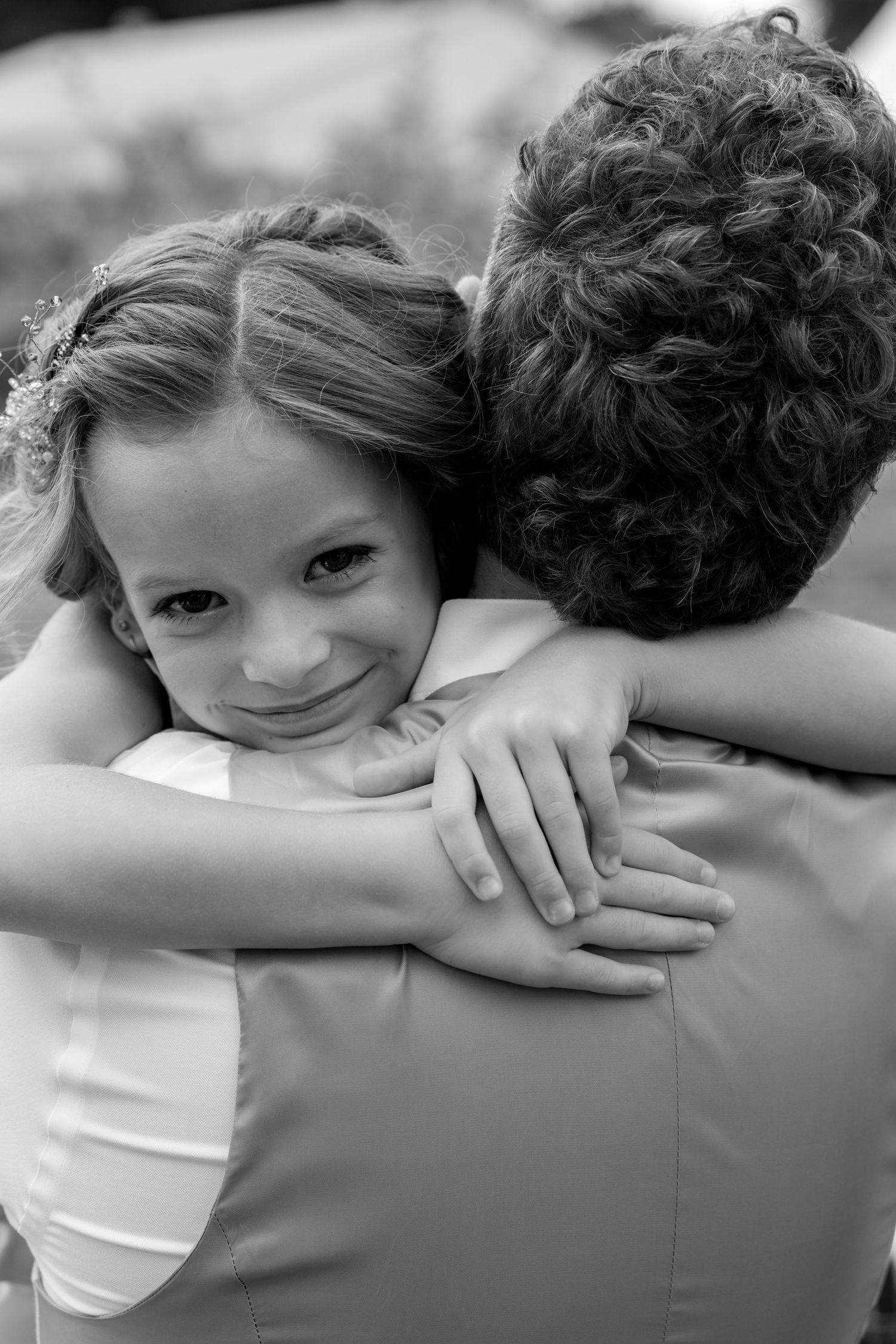 A smiling flower girl hugs the groom tightly during a wedding celebration in a black and white photo.