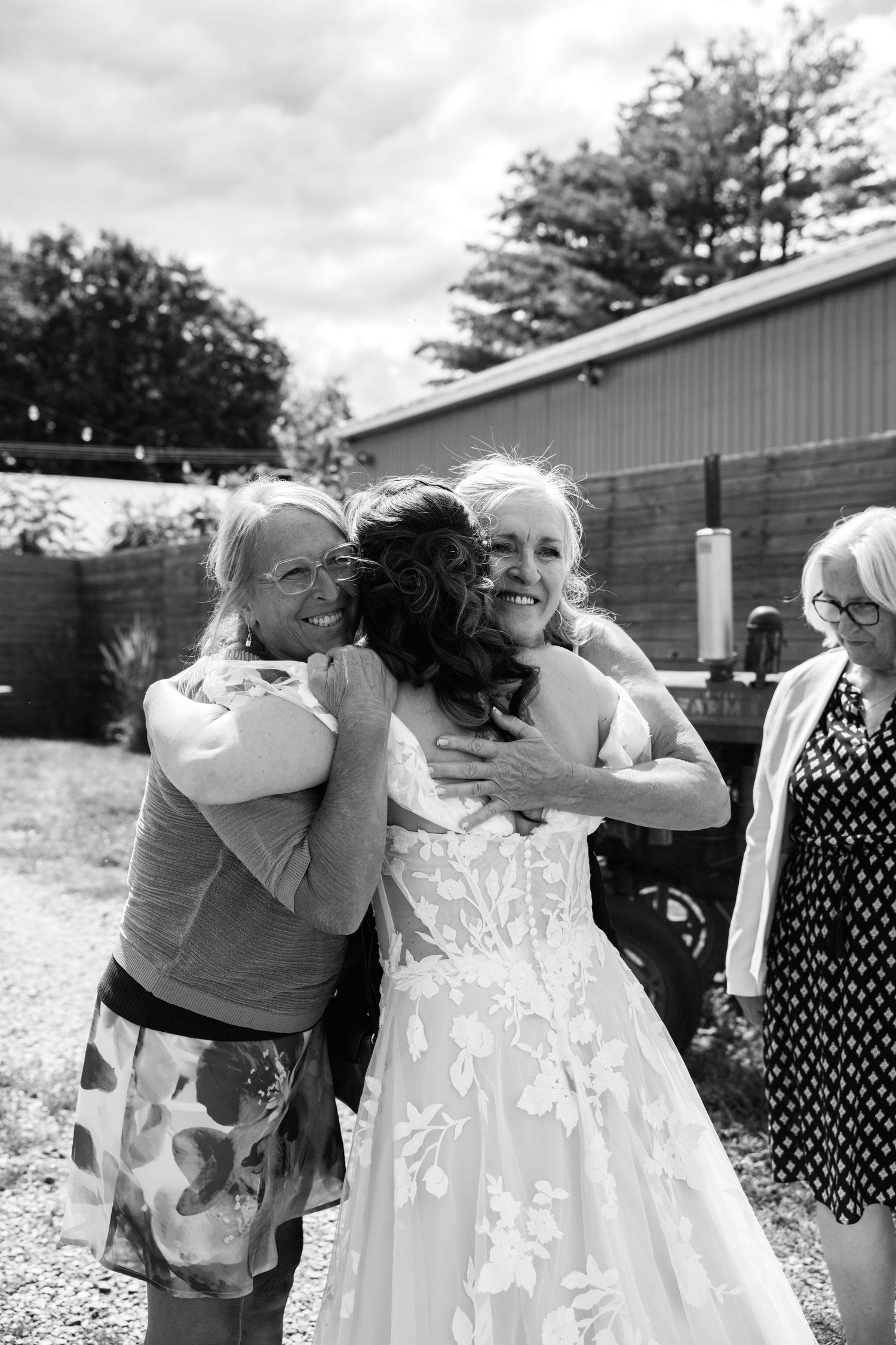 Bride hugged by two women guests outdoors in black and white photo.