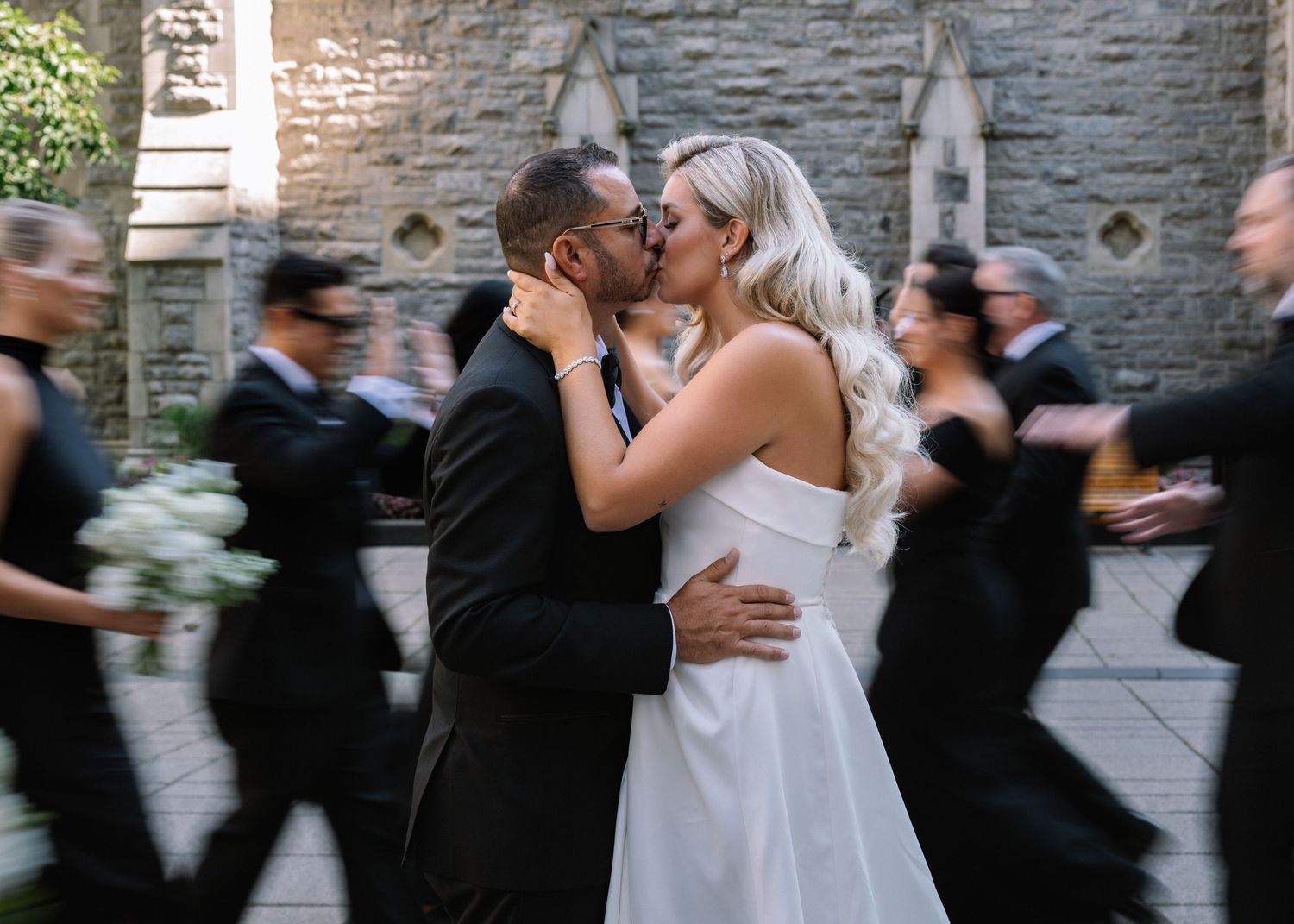 Newlyweds share a kiss while wedding party walks past a stone church.