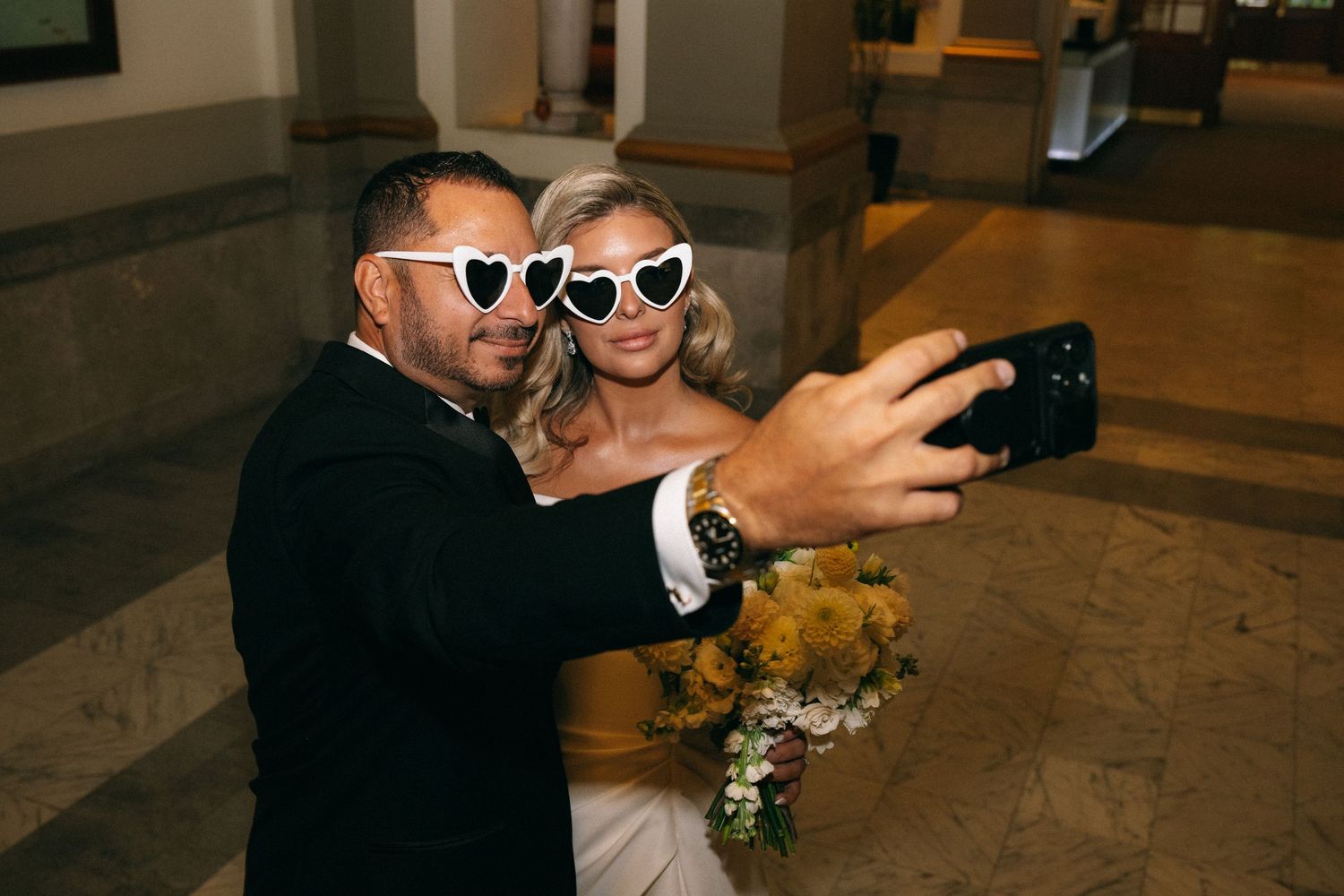 Bride in white dress and heart sunglasses takes selfie with groom in black suit holding yellow flower bouquet.
