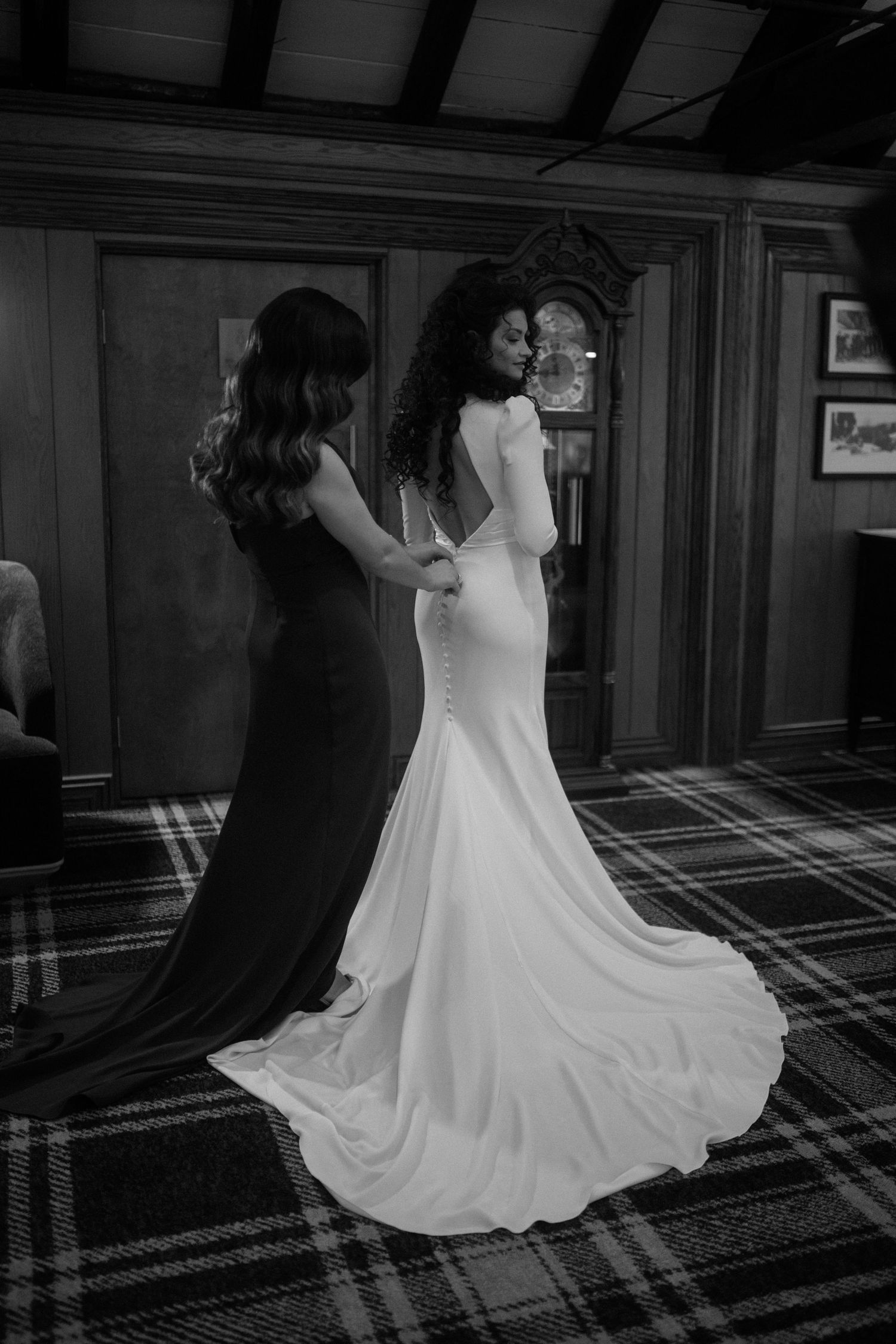 Black and white photo of a bride in a white gown being helped by a bridesmaid in a dark dress inside an elegant room.