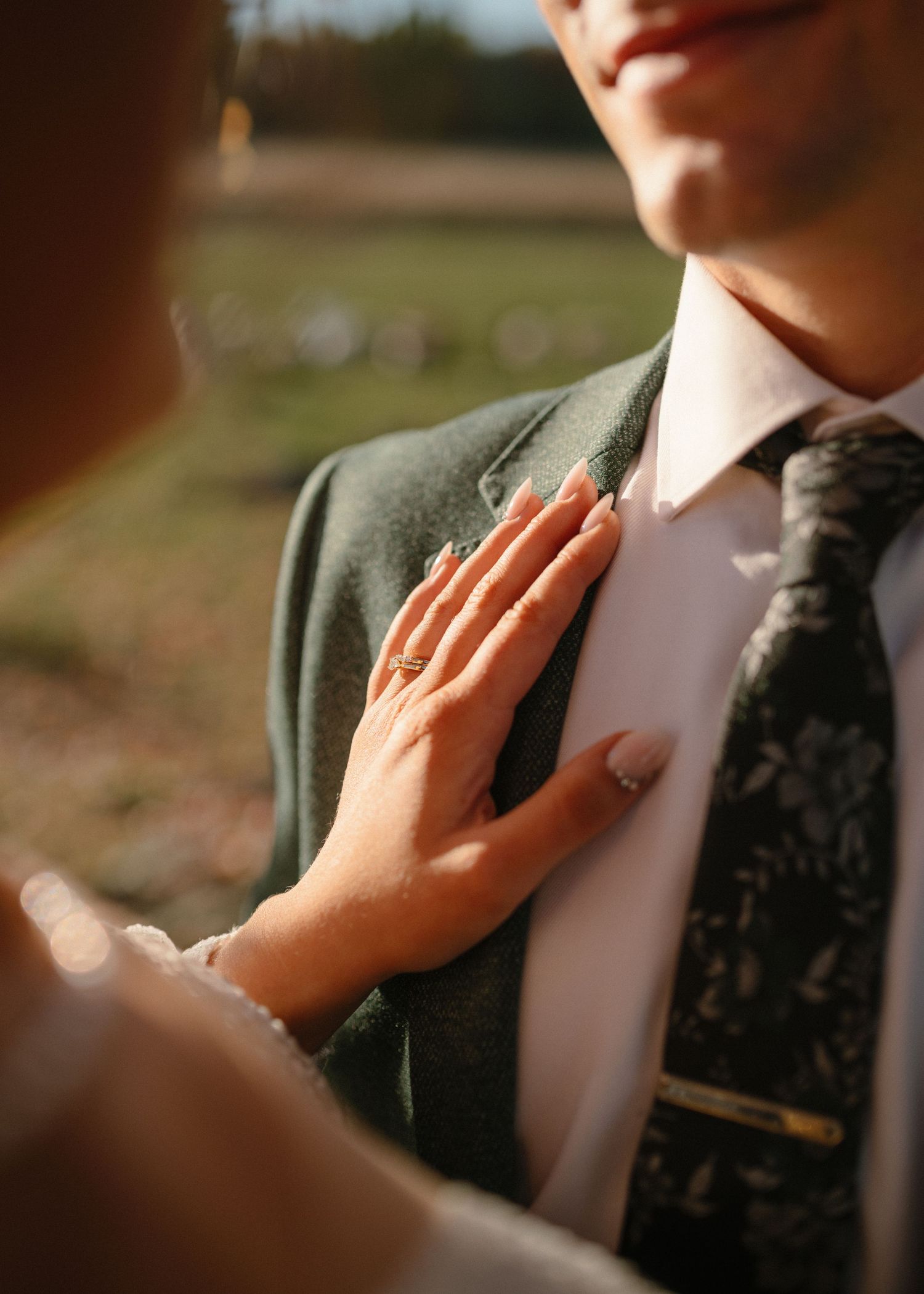 Bride placing her hand on grooms green suit chest, showing engagement ring in golden sunlight.