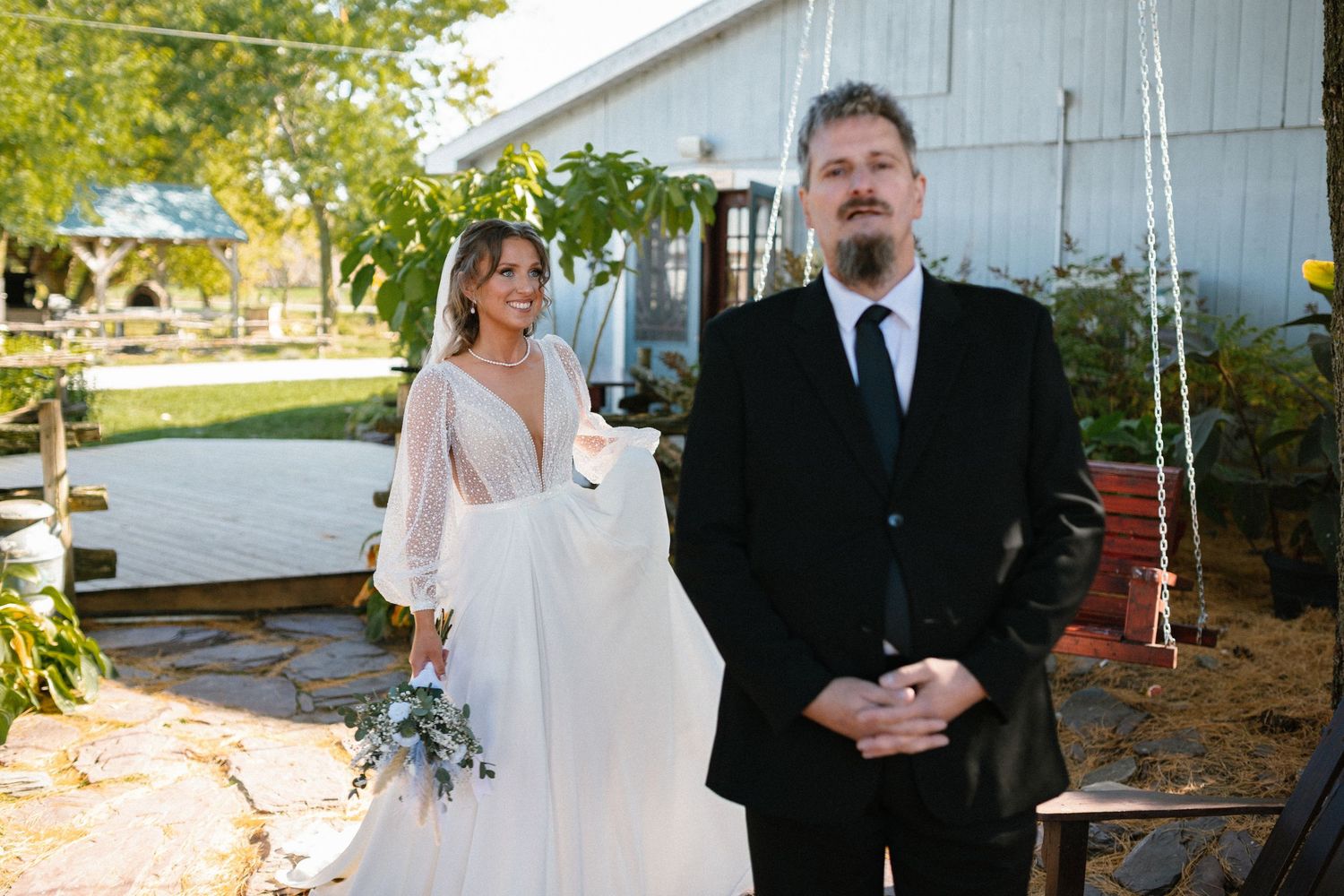 Bride in a sheer long-sleeve wedding gown holding bouquet stands with groom in black suit outdoors.