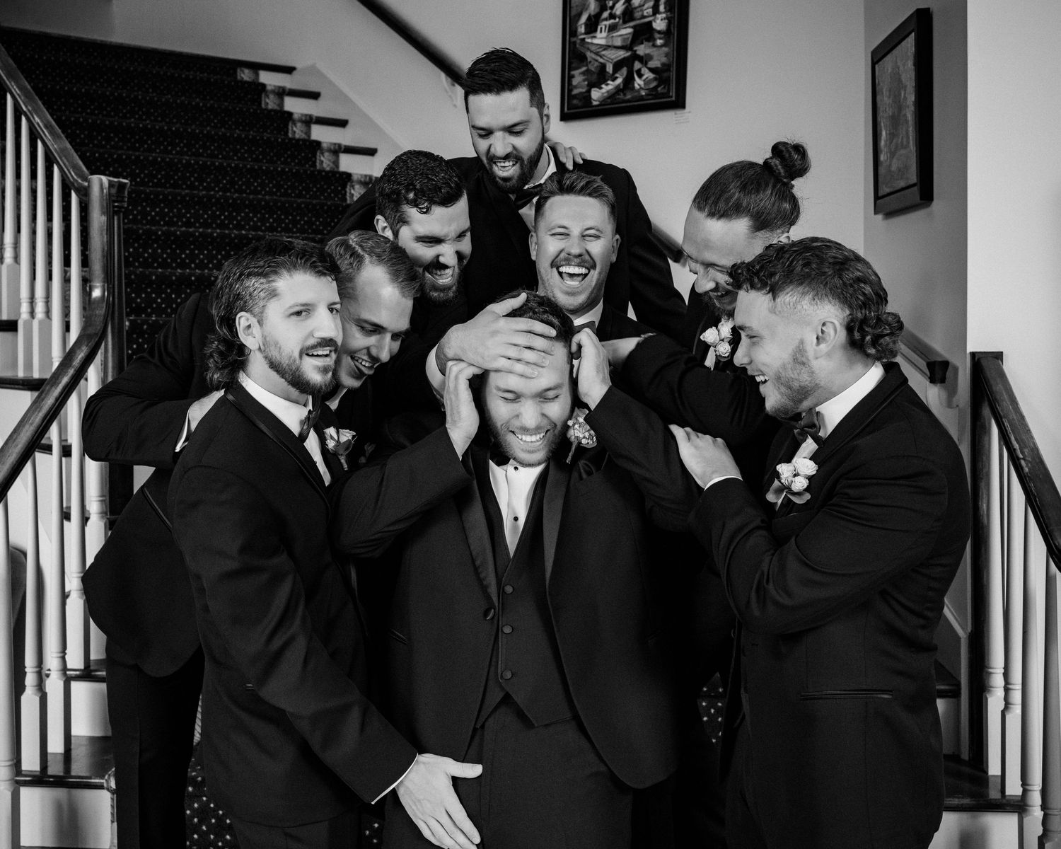 Groom surrounded by laughing groomsmen in black suits on staircase in black and white wedding photo.