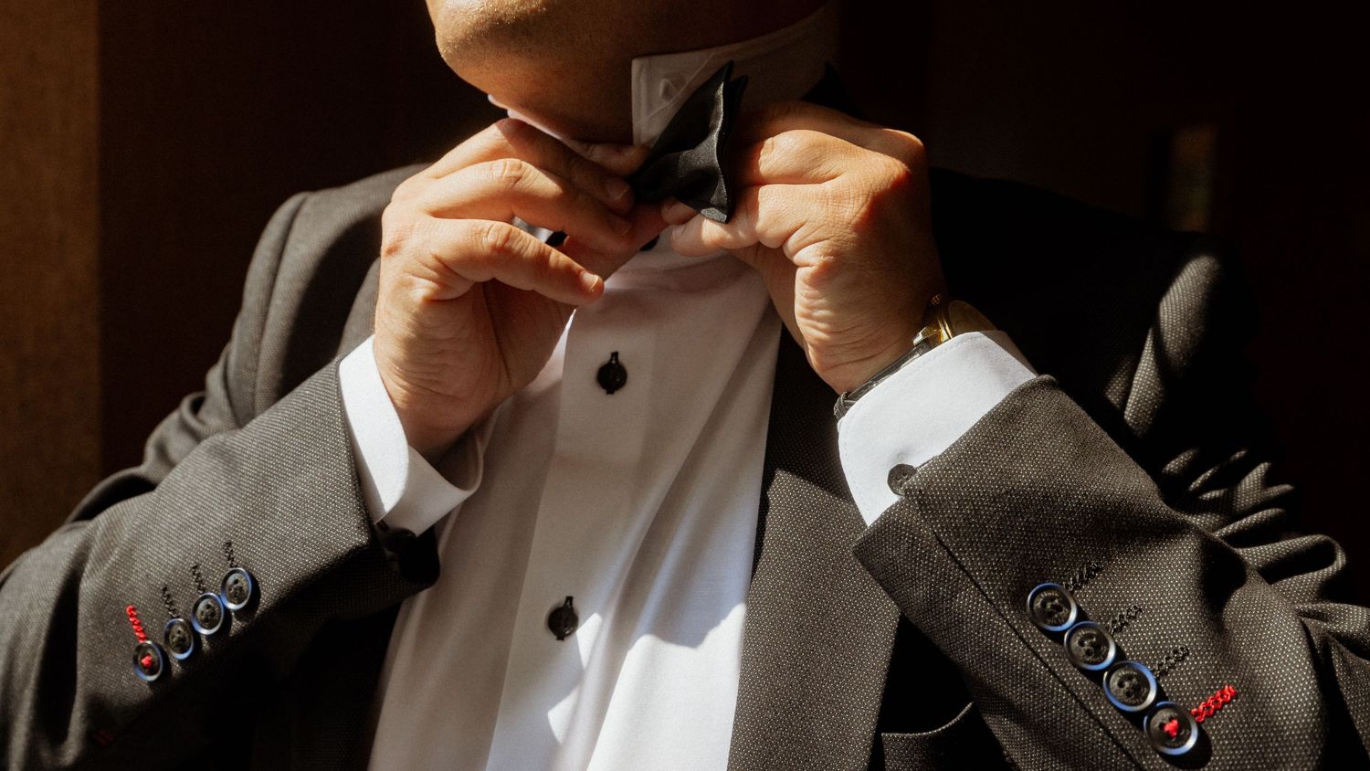 A man in a tuxedo adjusting his black bow tie, dressed in formal attire with a white dress shirt.