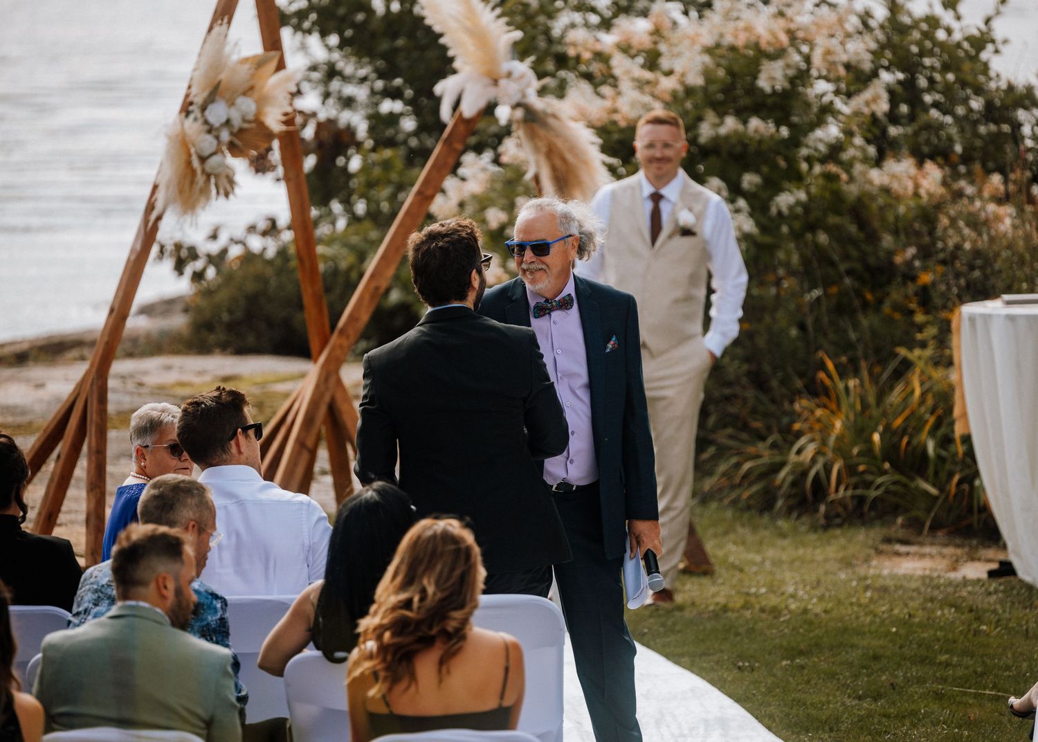 Elderly man in sunglasses greets groom at outdoor waterfront wedding ceremony with boho pampas grass arch.