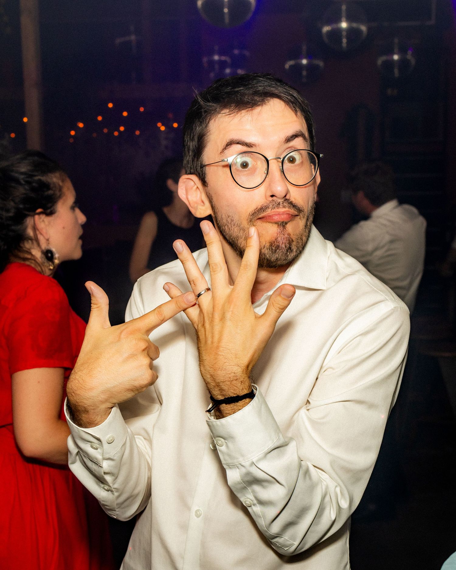 Man with glasses making funny face and hand gestures at a party, wearing white shirt with woman in red dress behind him.