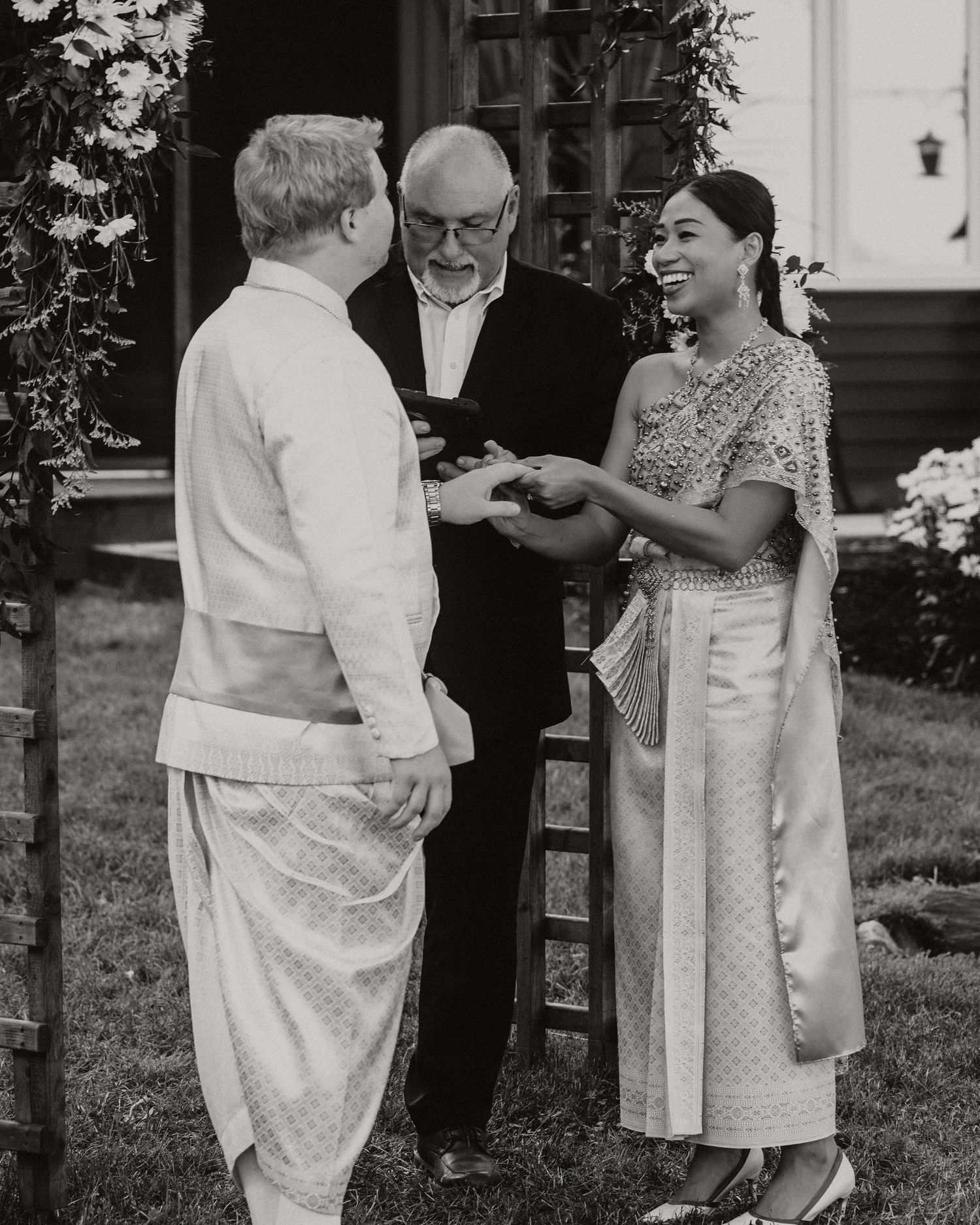 Couple exchanging rings during outdoor wedding ceremony with officiant, bride in traditional Thai dress, black and white photo.