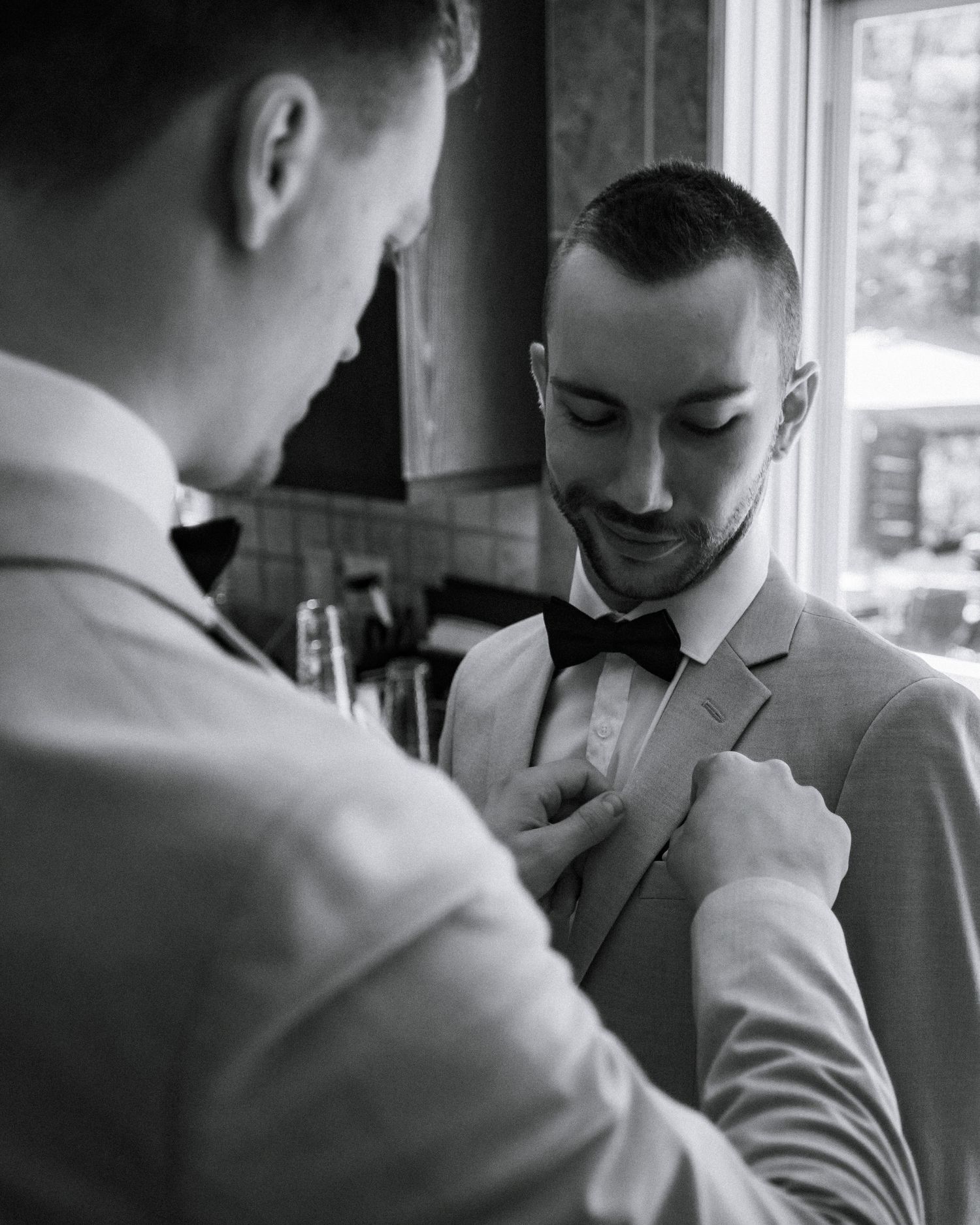 Groom smiling as groomsman adjusts his bow tie in black and white wedding preparation photo.