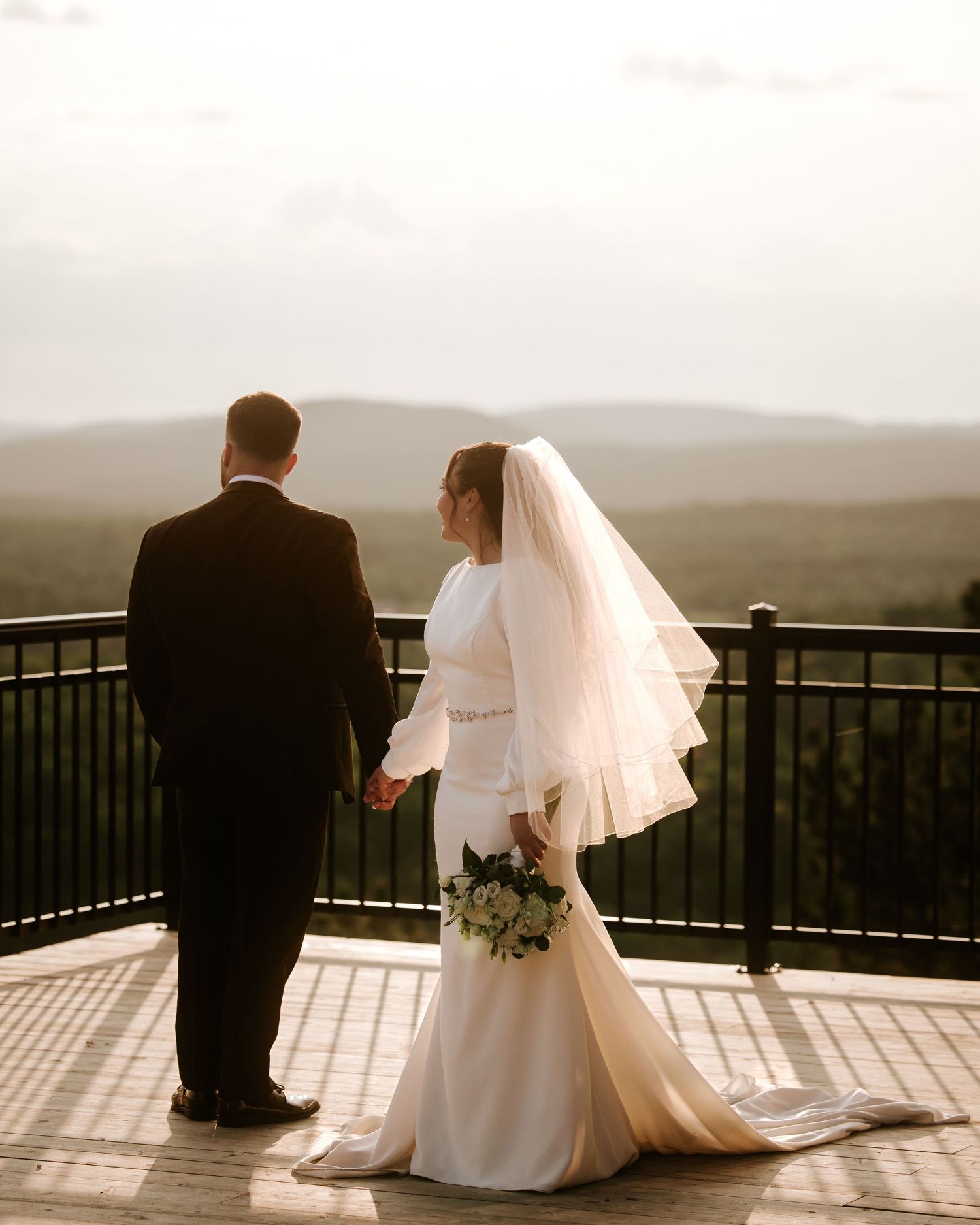 Bride and groom holding hands on scenic overlook deck with mountain views at sunset.