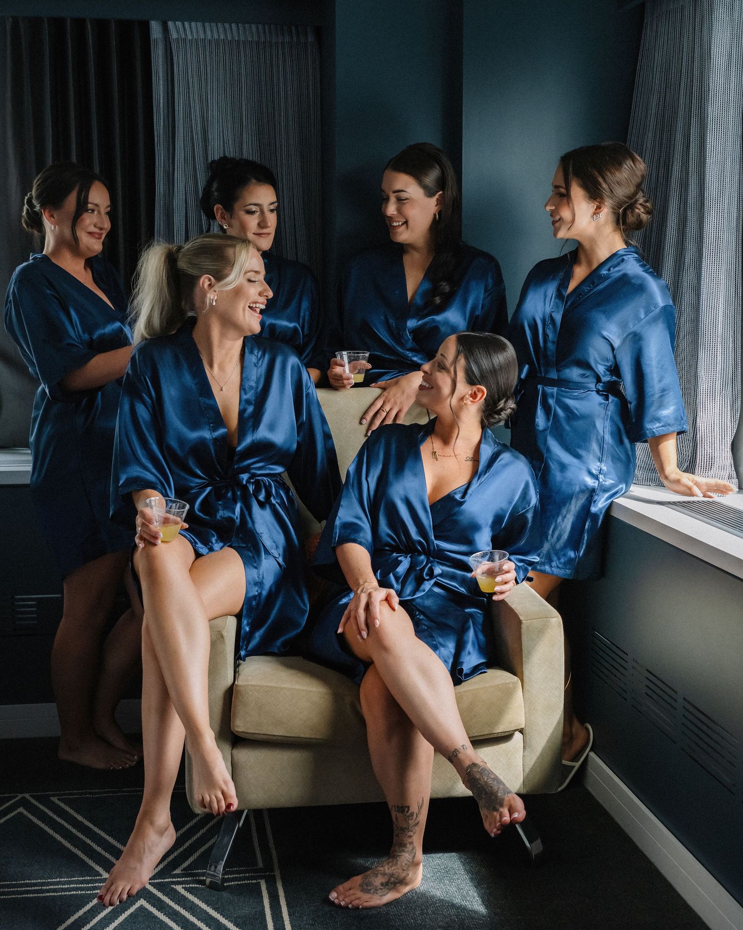 Bridesmaids in matching blue satin robes gathered together laughing and celebrating before wedding ceremony.