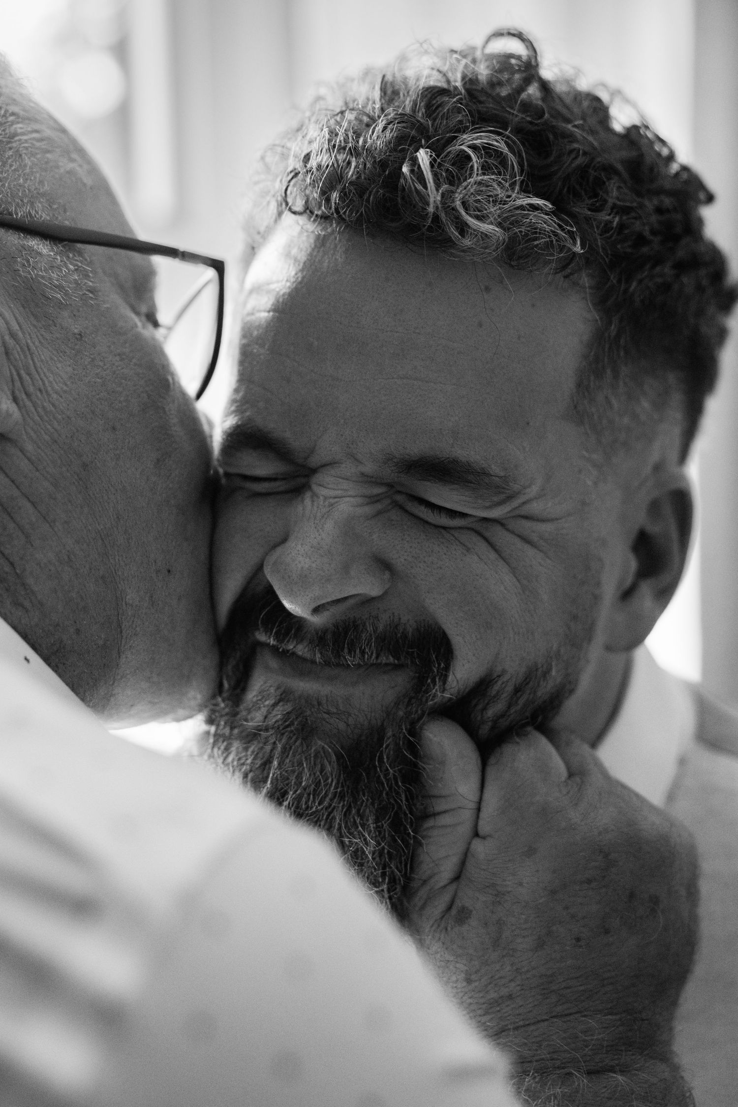 A bearded man with curly hair smiles warmly while receiving an affectionate kiss on the cheek from an older person.