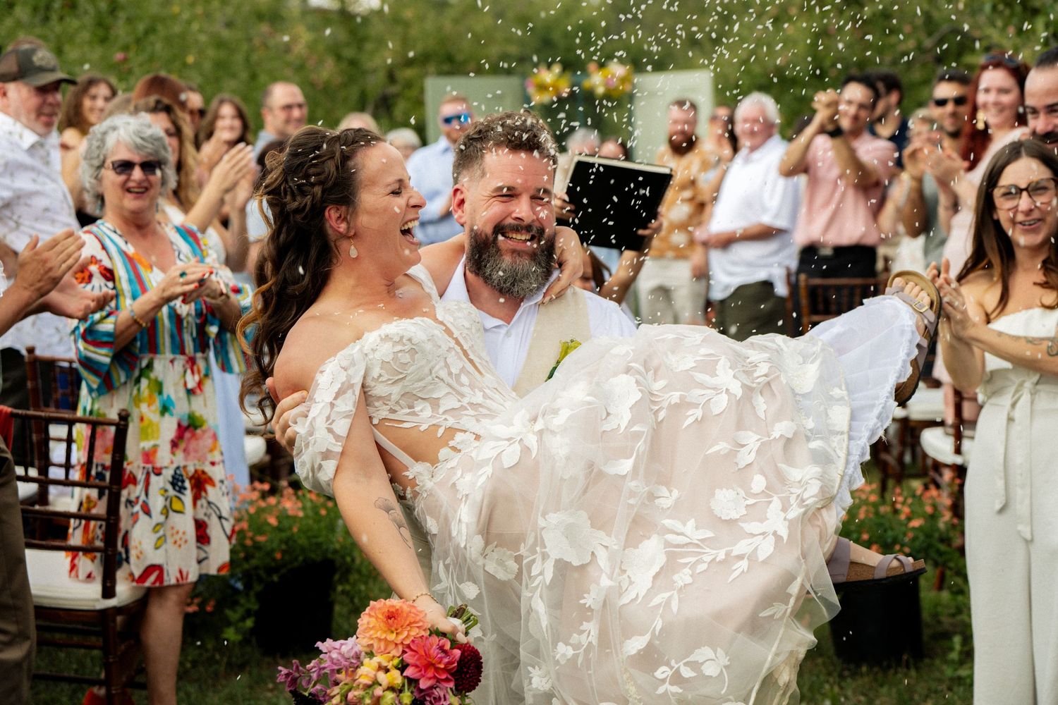 Smiling groom carries his bride in a floral wedding dress as guests toss confetti outdoors.
