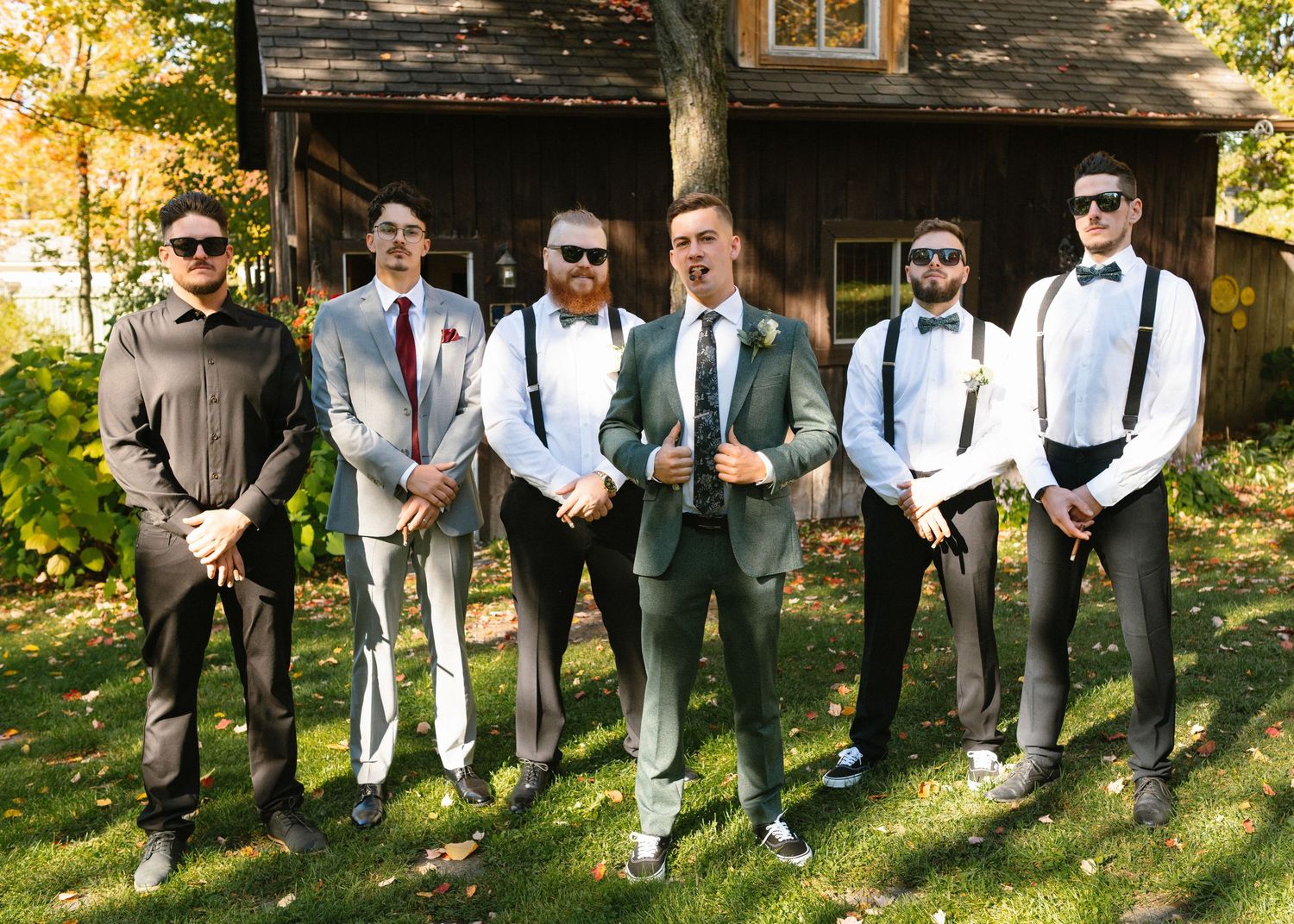 Groom and groomsmen in suspenders and bow ties posing outdoors near rustic barn on wedding day.