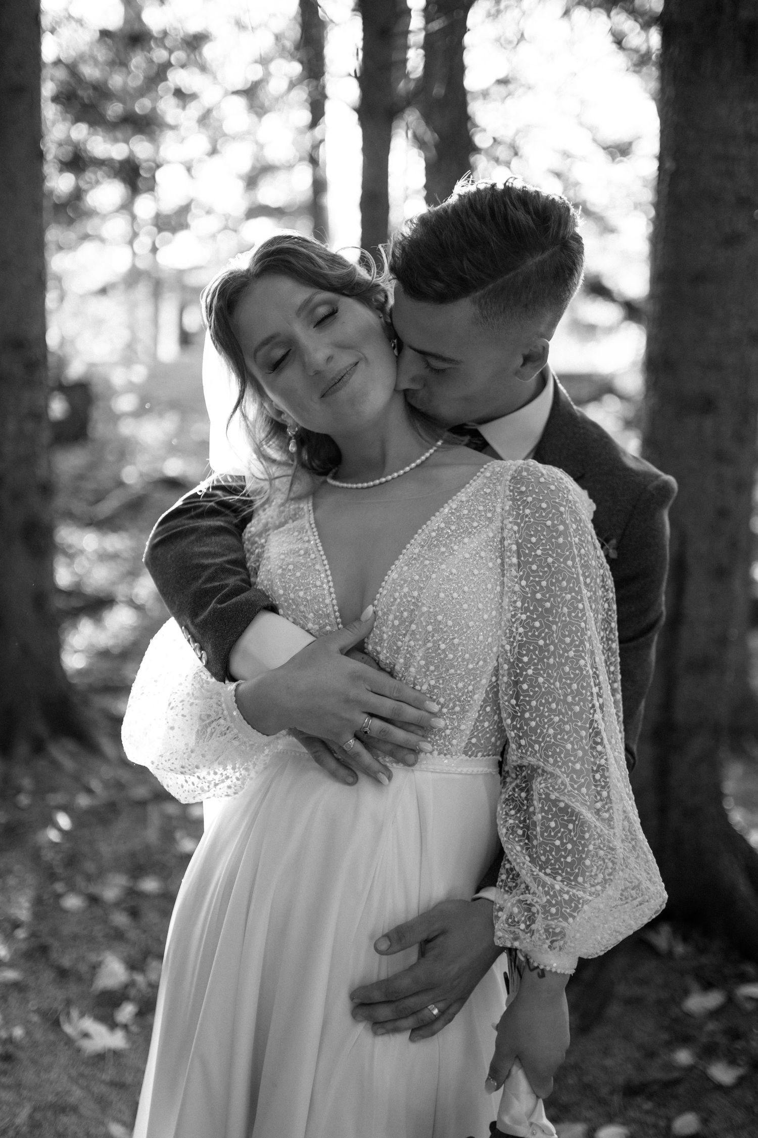 Black and white wedding photo of groom kissing bride on cheek while embracing in a forest setting.