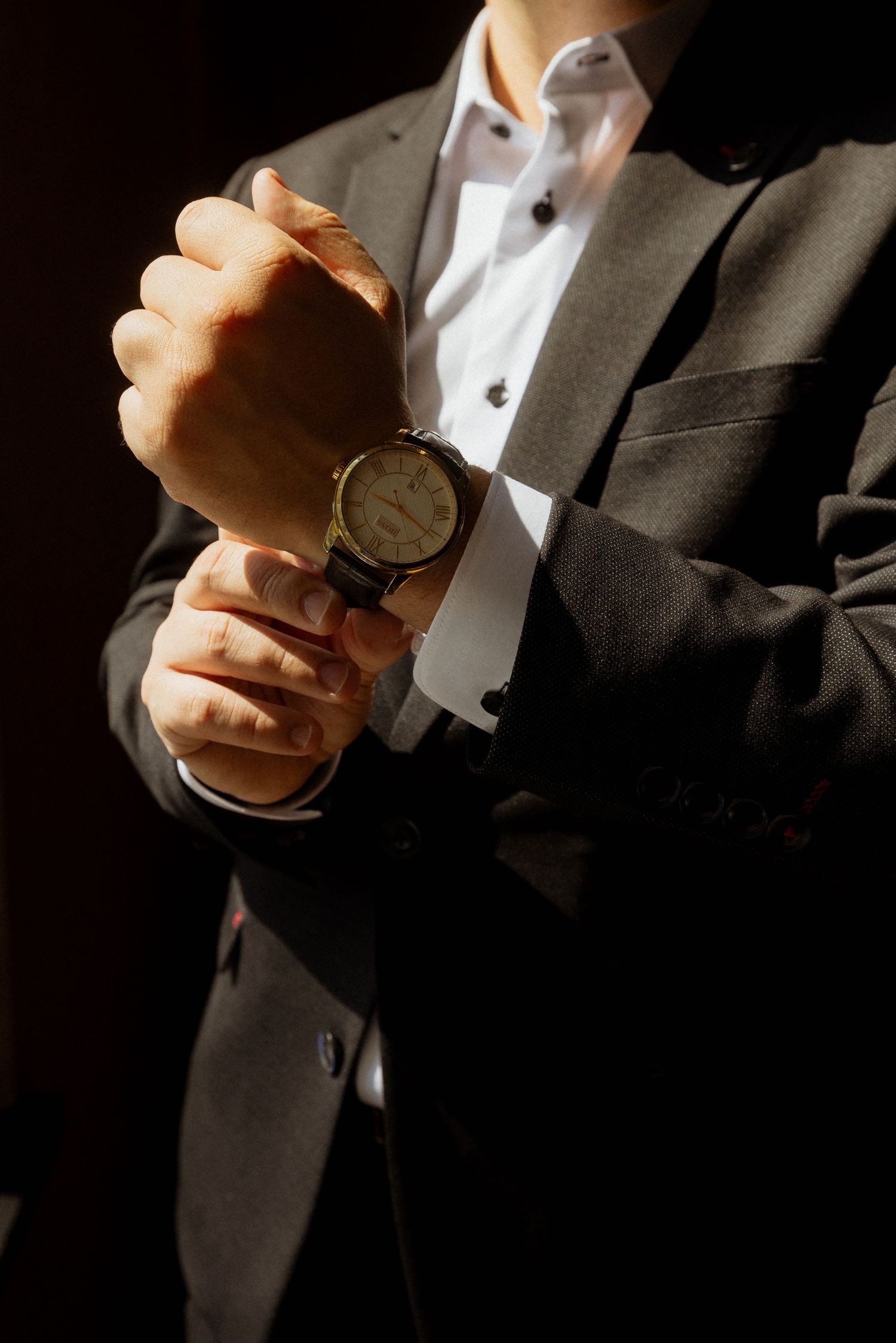Man in dark suit adjusting a classic leather wristwatch in dramatic lighting.