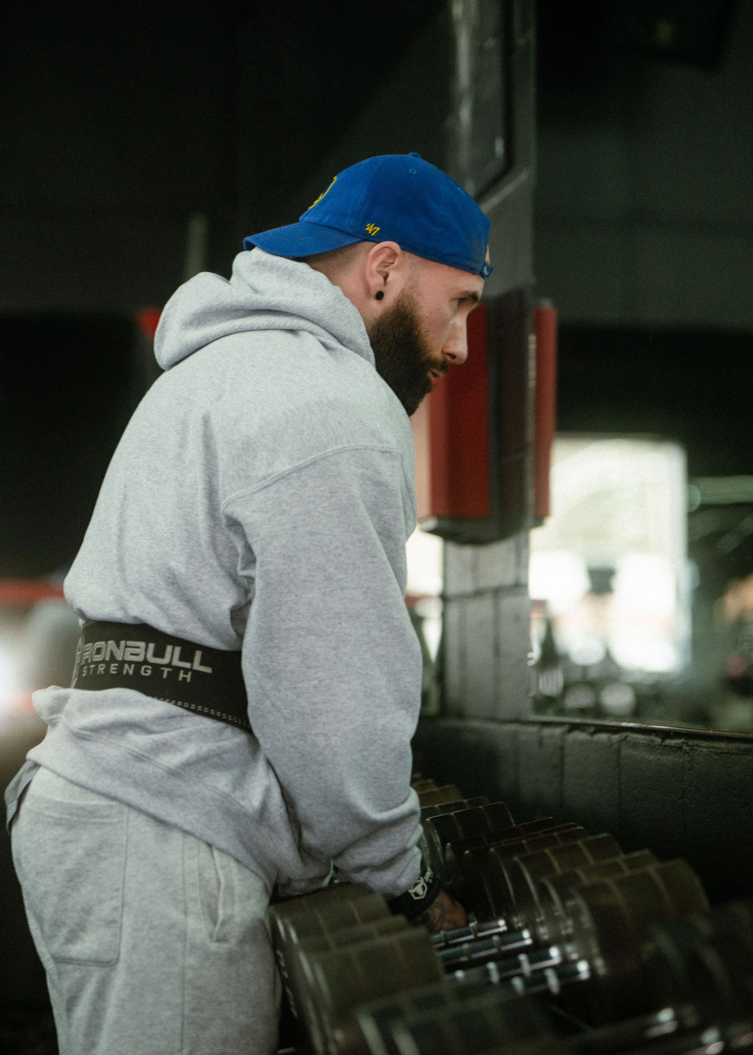 Man in grey hoodie and blue cap selecting weights at a dumbbell rack in a dark gym.