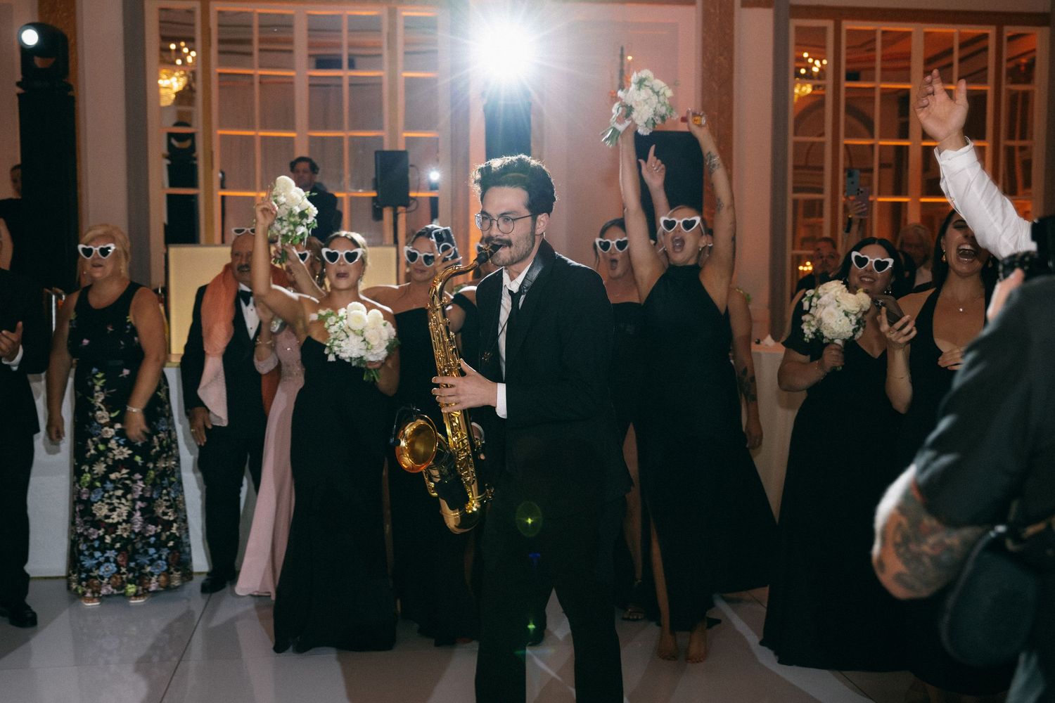 Saxophonist performing at wedding reception while bridesmaids in sunglasses dance and hold bouquets.