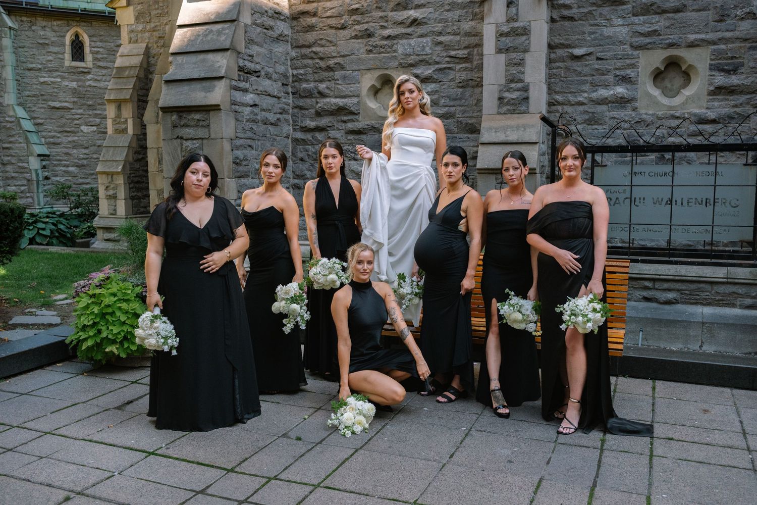 Bride posing with seven bridesmaids in black gowns holding white bouquets outside stone church.
