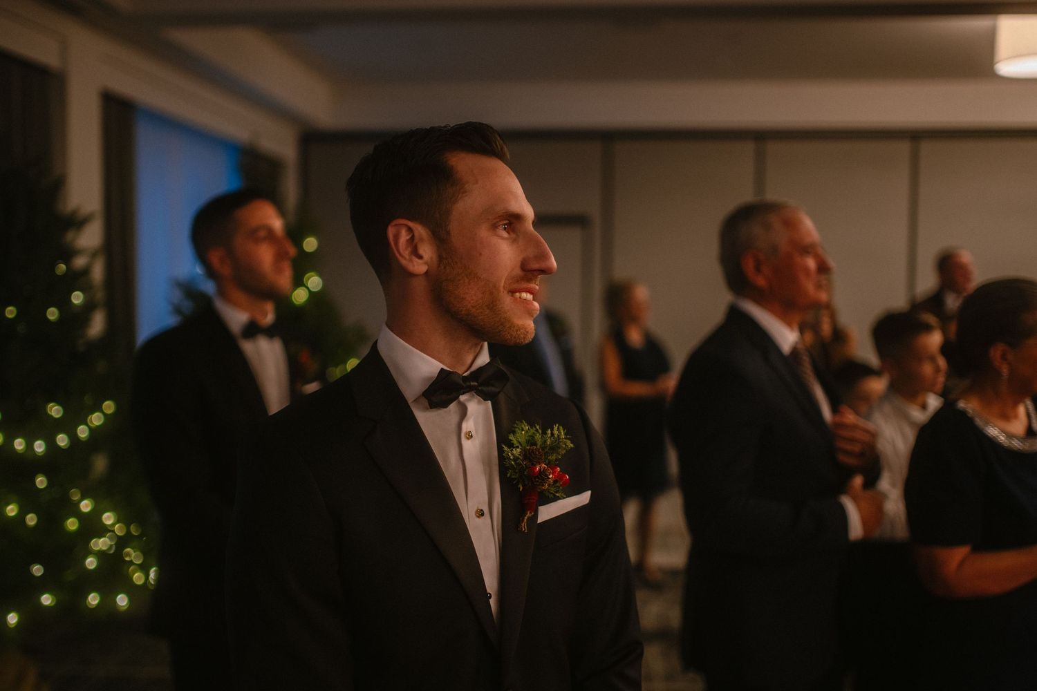 Groom in black tuxedo with floral boutonniere stands at wedding reception with guests and Christmas tree in background.