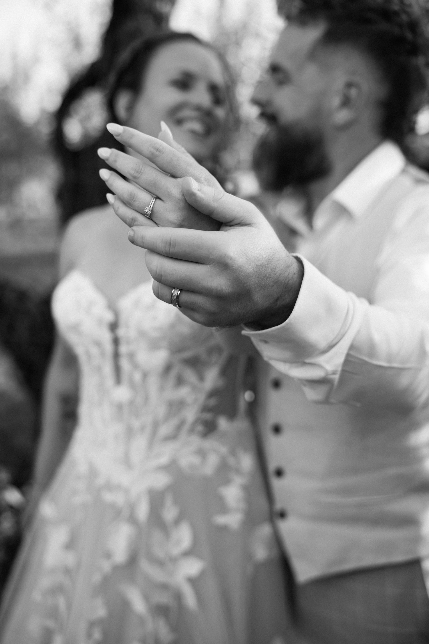 Black and white photo of smiling bride and groom showing off wedding rings outdoors.