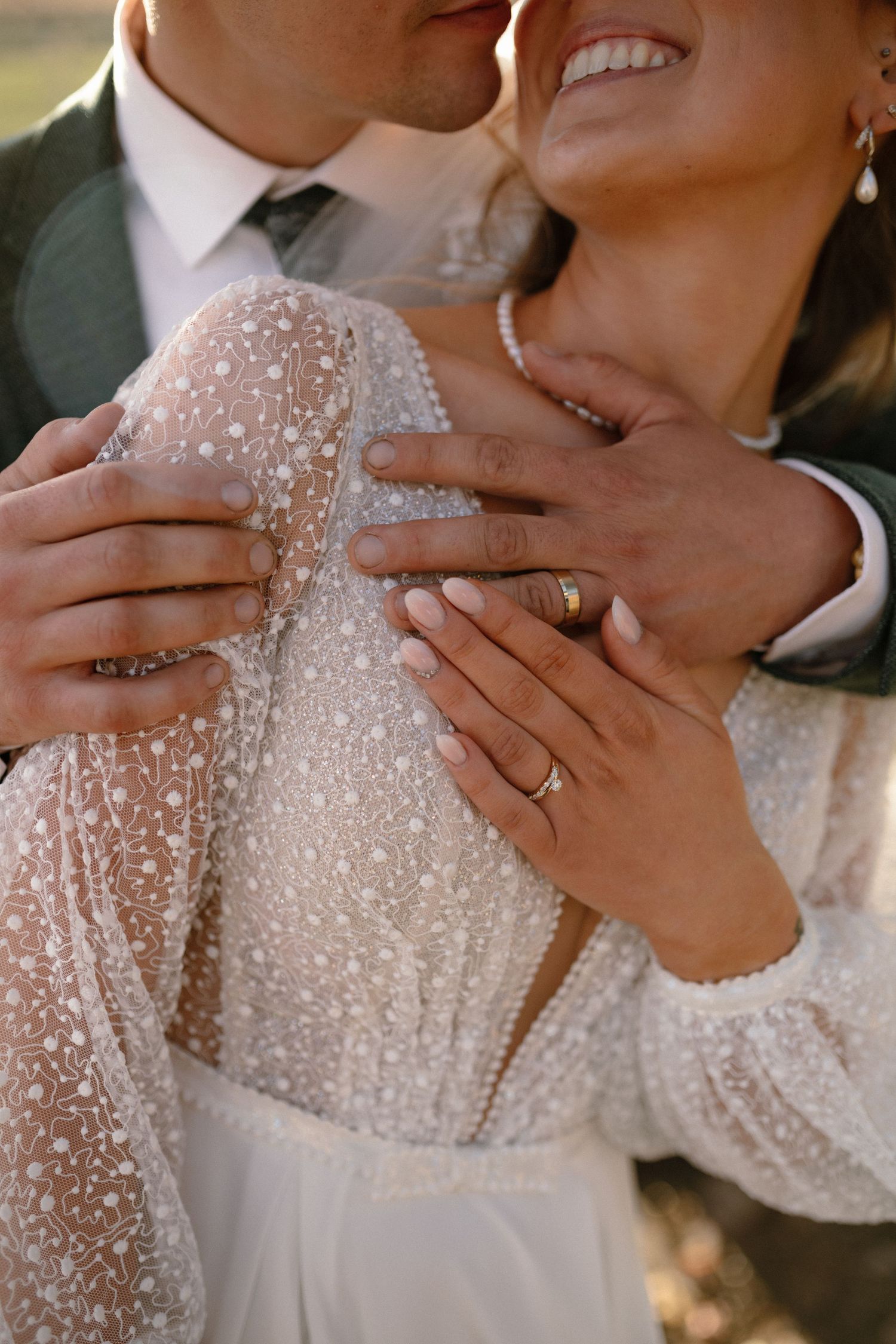 Groom embracing bride in a beaded lace wedding dress, both wearing gold wedding rings.