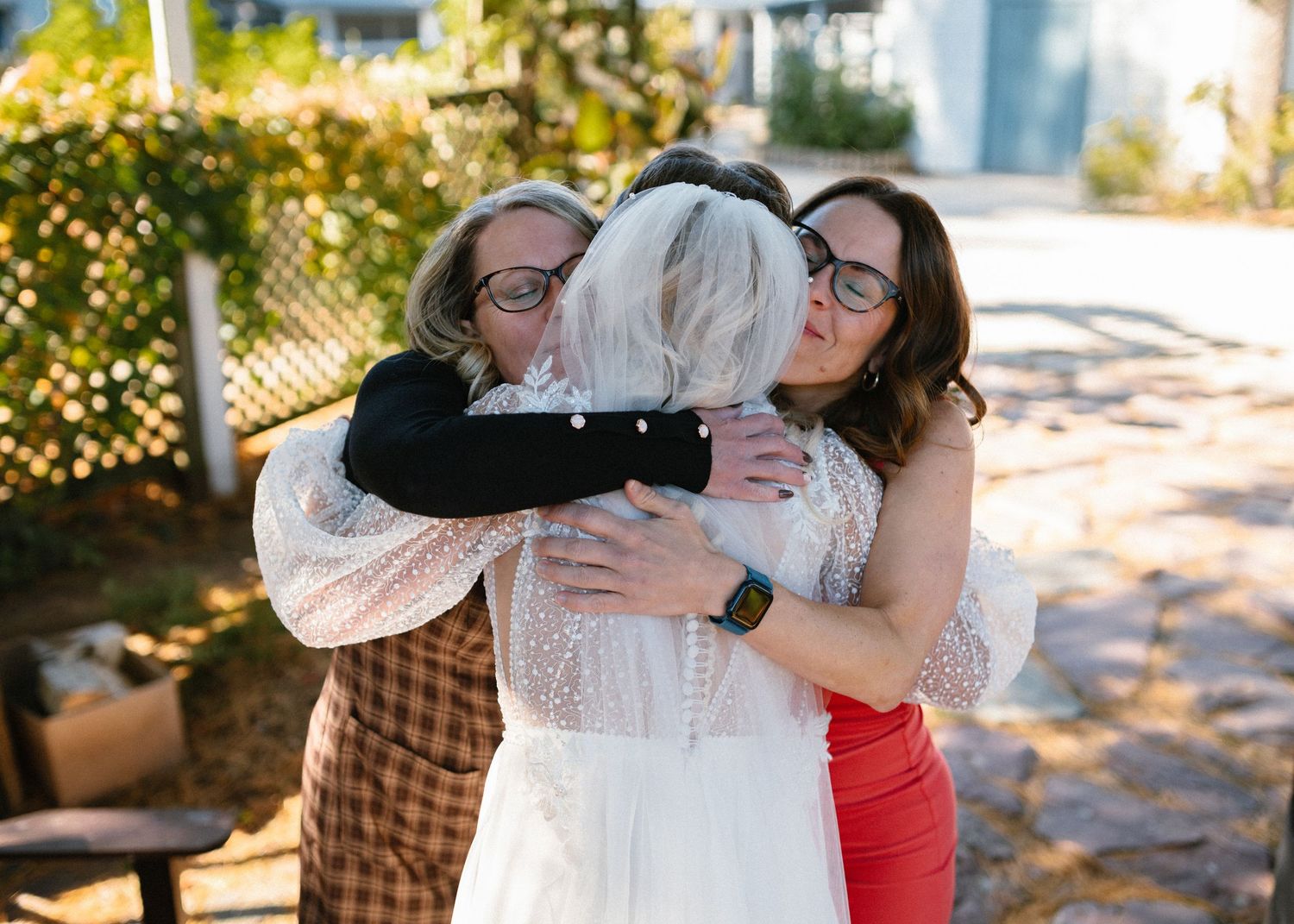 Bride in white lace gown sharing a group hug with two women guests outdoors.
