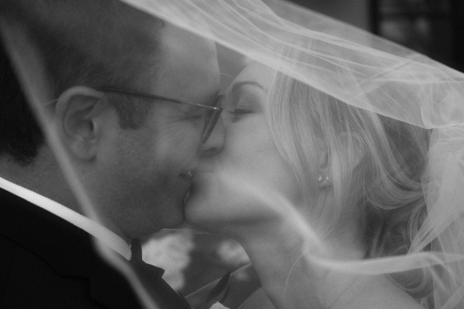 Intimate black and white portrait of bride and groom kissing beneath sheer bridal veil on wedding day.