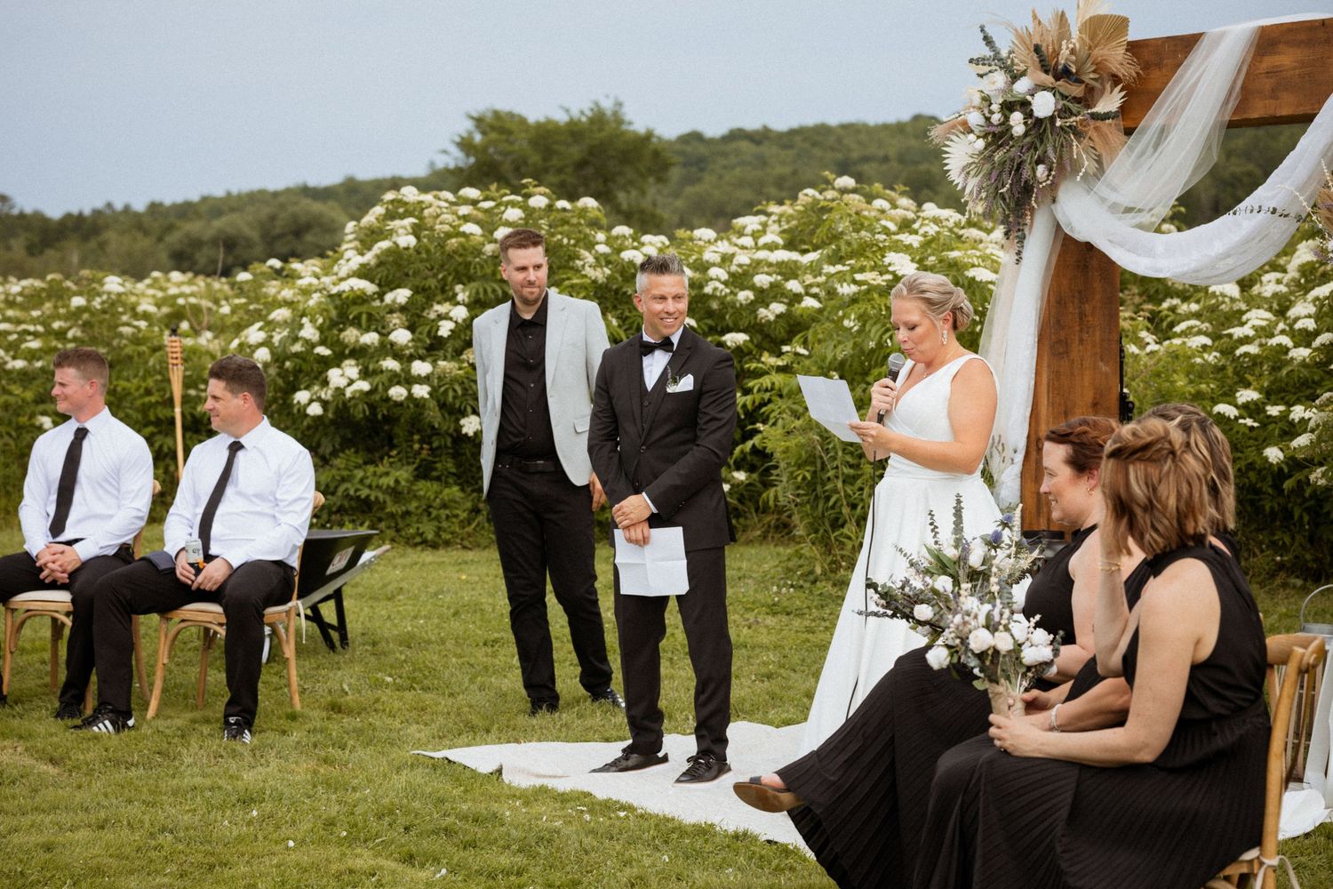 Outdoor wedding ceremony on green lawn with groom in tuxedo, officiant, groomsmen, and bridesmaids in black dresses.