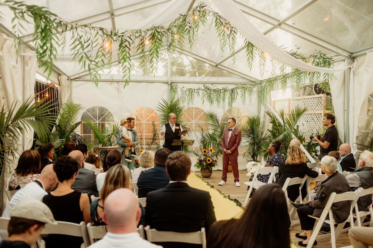 Elegant outdoor wedding ceremony under a white tent adorned with lush greenery, guests seated in white chairs.