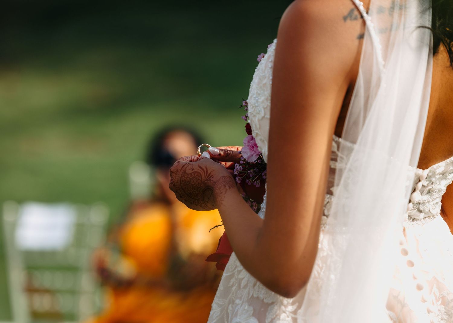 Bride in white lace gown holding a small bouquet during an outdoor wedding ceremony with guests seated in background.