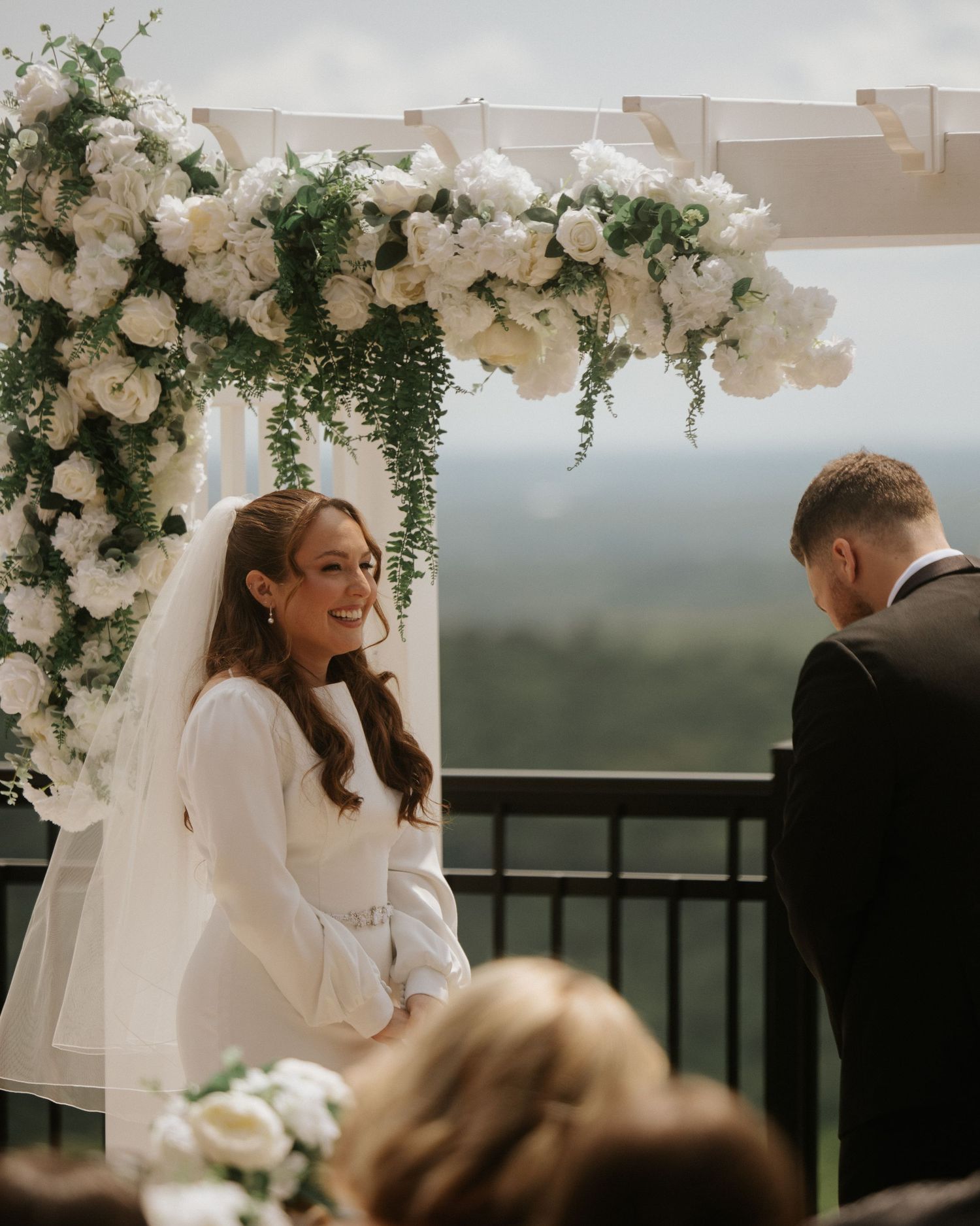 Bride smiling under white floral arch with greenery during outdoor wedding ceremony with scenic views.