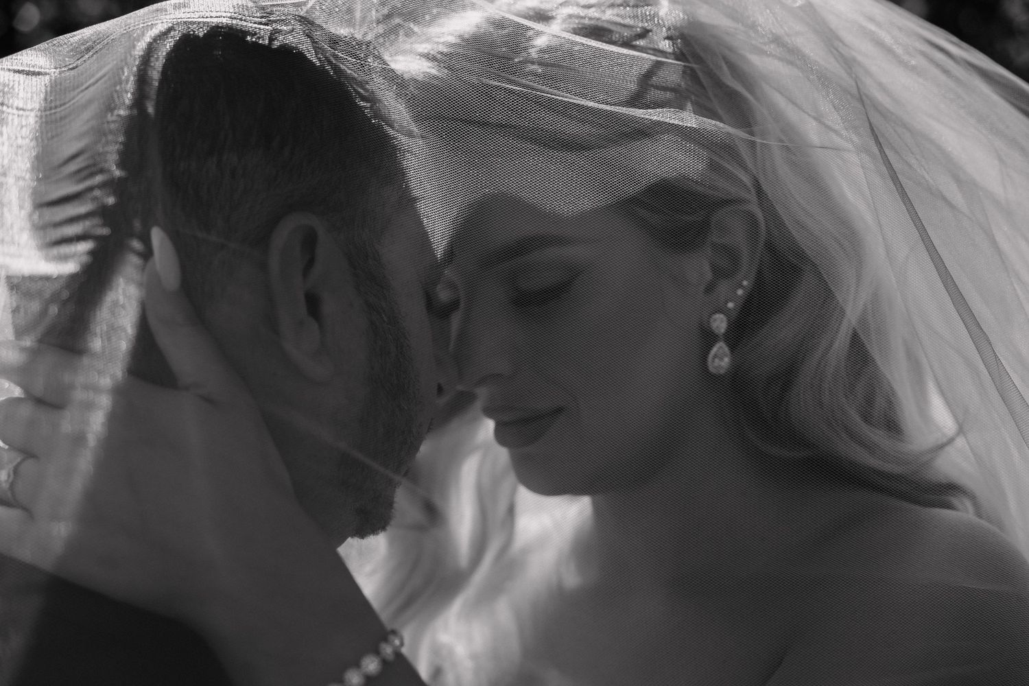 Bride and groom sharing intimate moment under wedding veil in black and white.