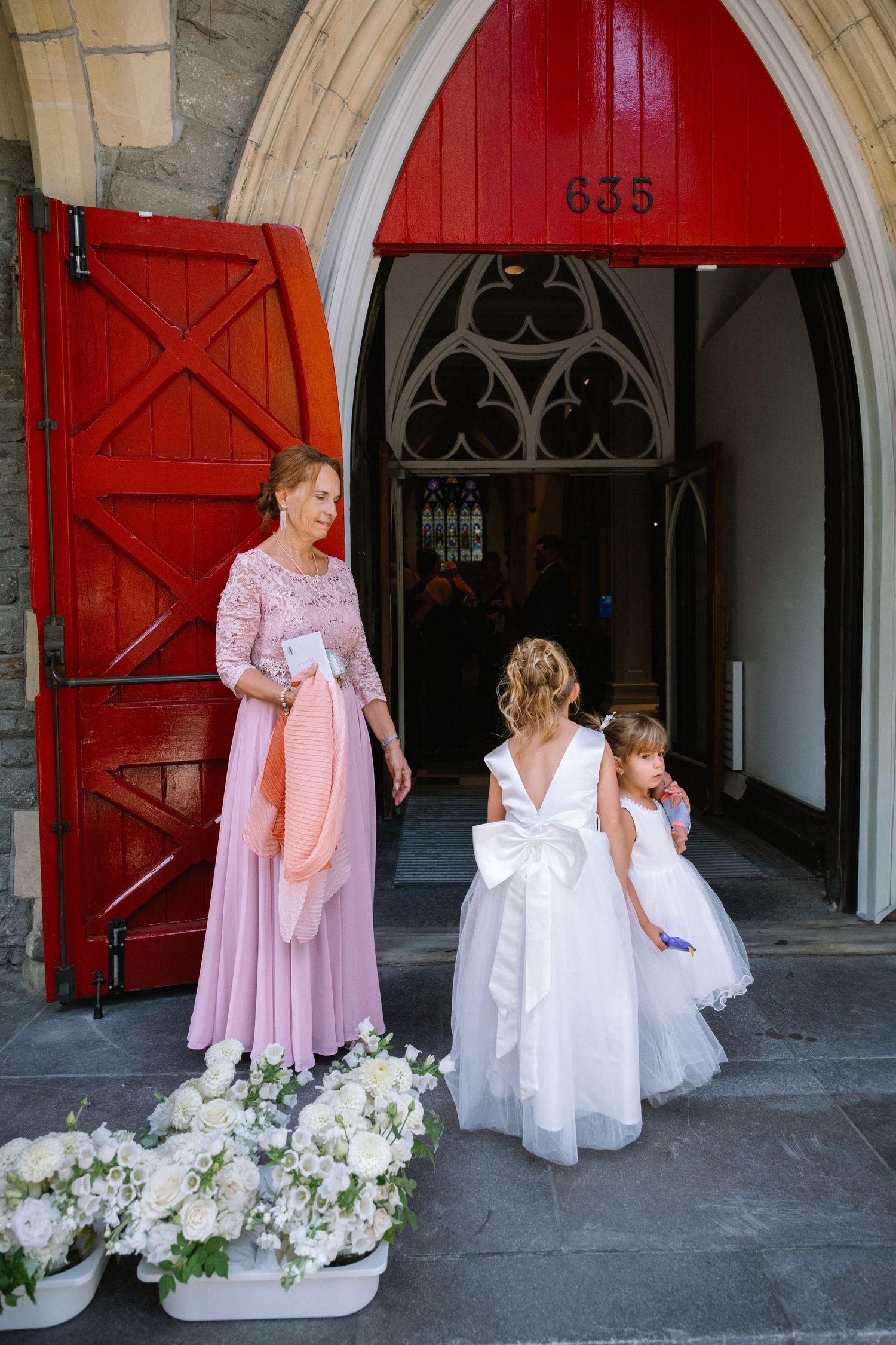 Woman in pink dress assists two flower girls in white outside church with red doors numbered 635.