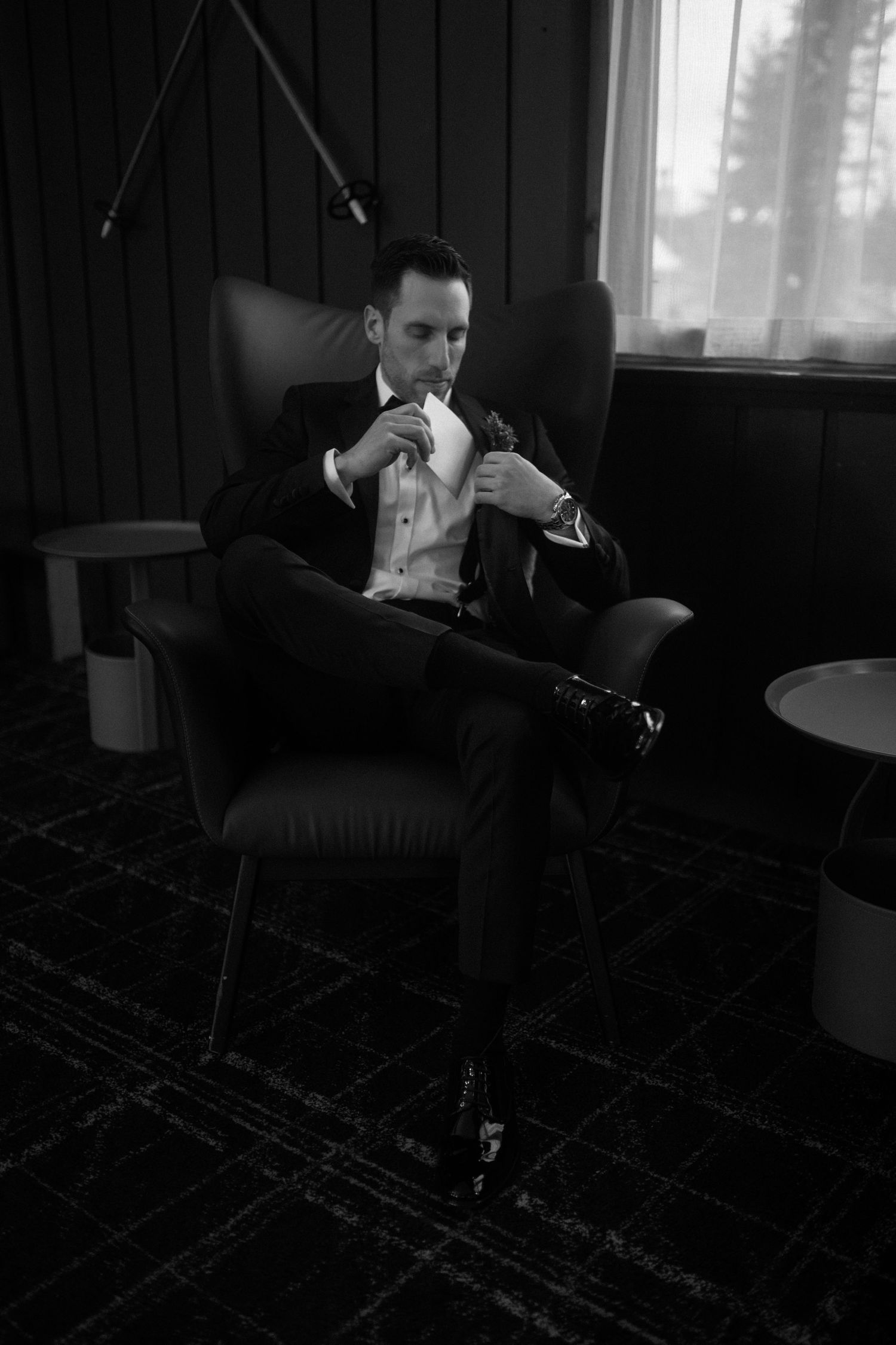 Groom in black suit adjusting his tie while seated in a leather chair in an elegant hotel room.