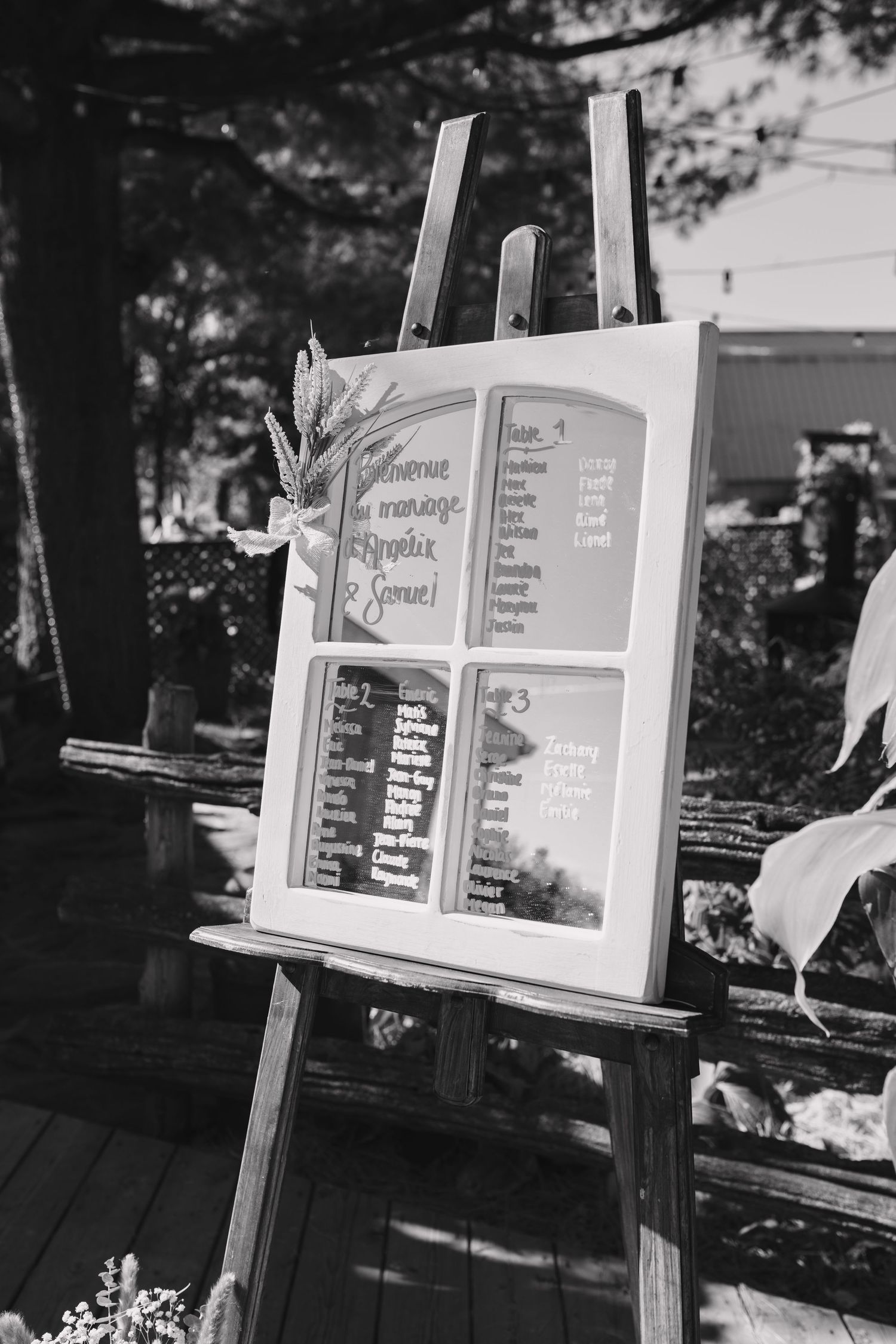Black and white photo of a wedding seating chart displayed on an easel outdoors.