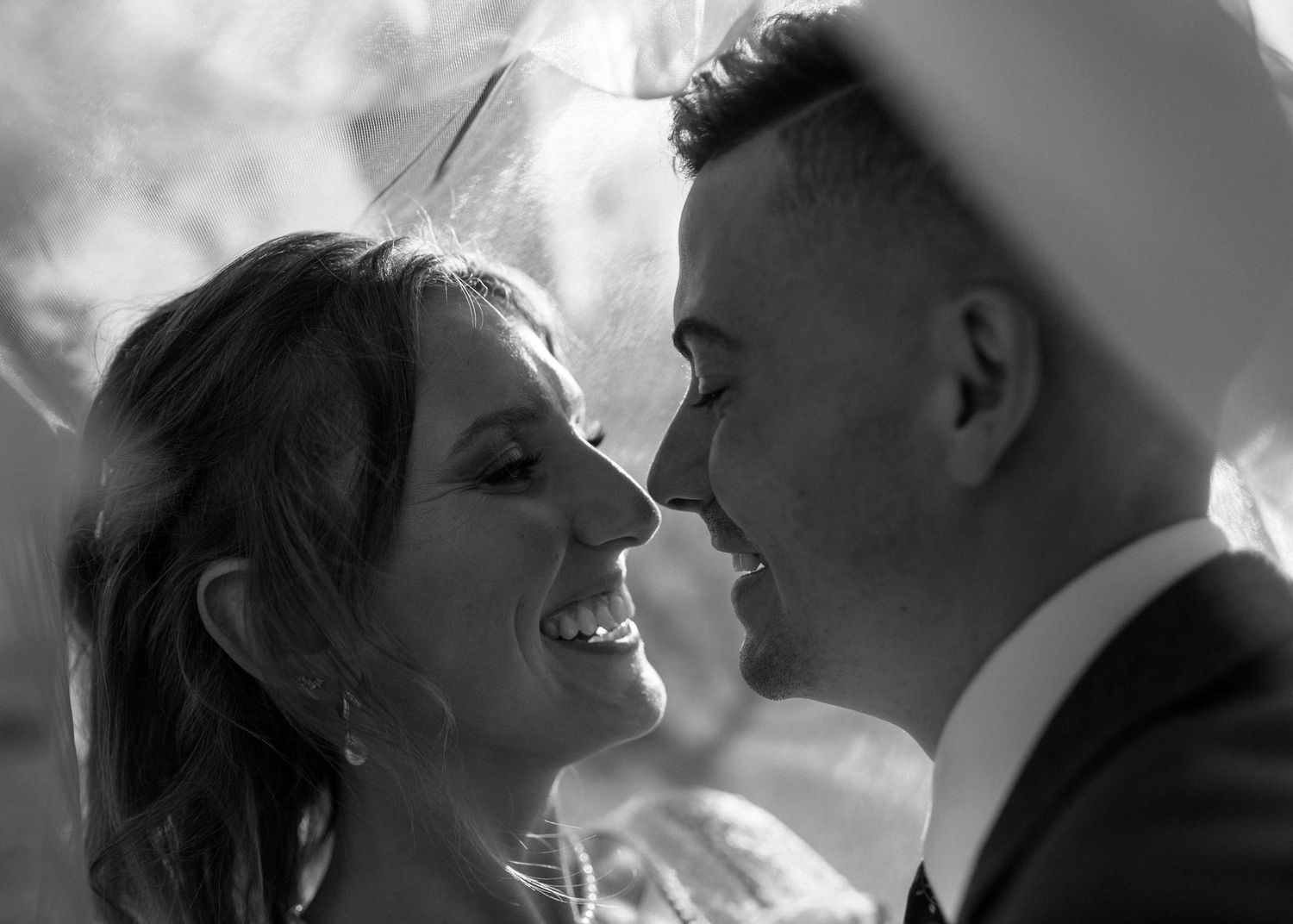 Close-up black and white portrait of smiling bride and groom gazing at each other under veil.