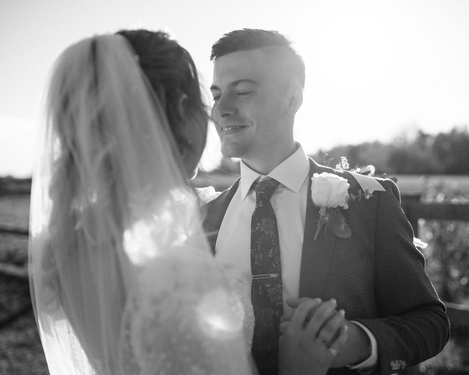 Black and white photo of a smiling groom gazing at his bride in a veil outdoors.