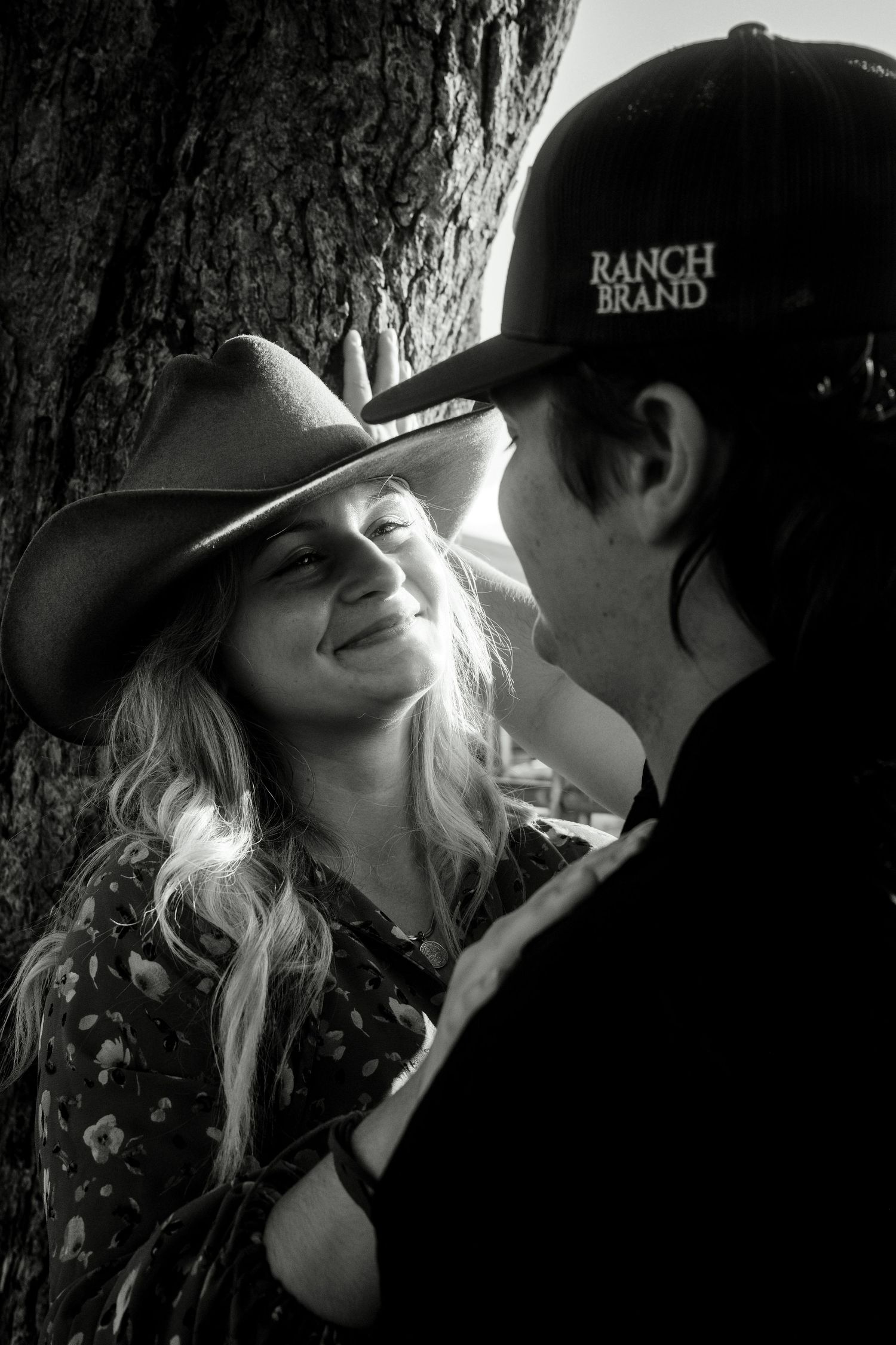 Black and white romantic portrait of smiling woman in cowboy hat with man wearing Ranch Brand cap by tree.