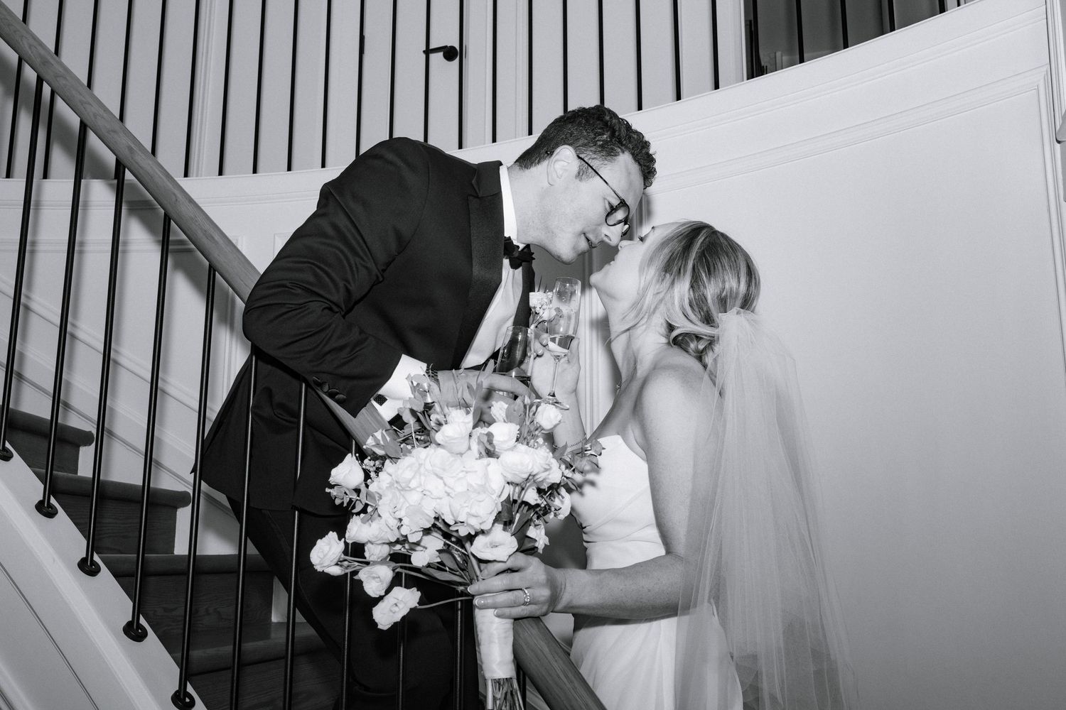 Bride and groom share tender kiss on staircase, bride holding white rose bouquet in black and white photo.