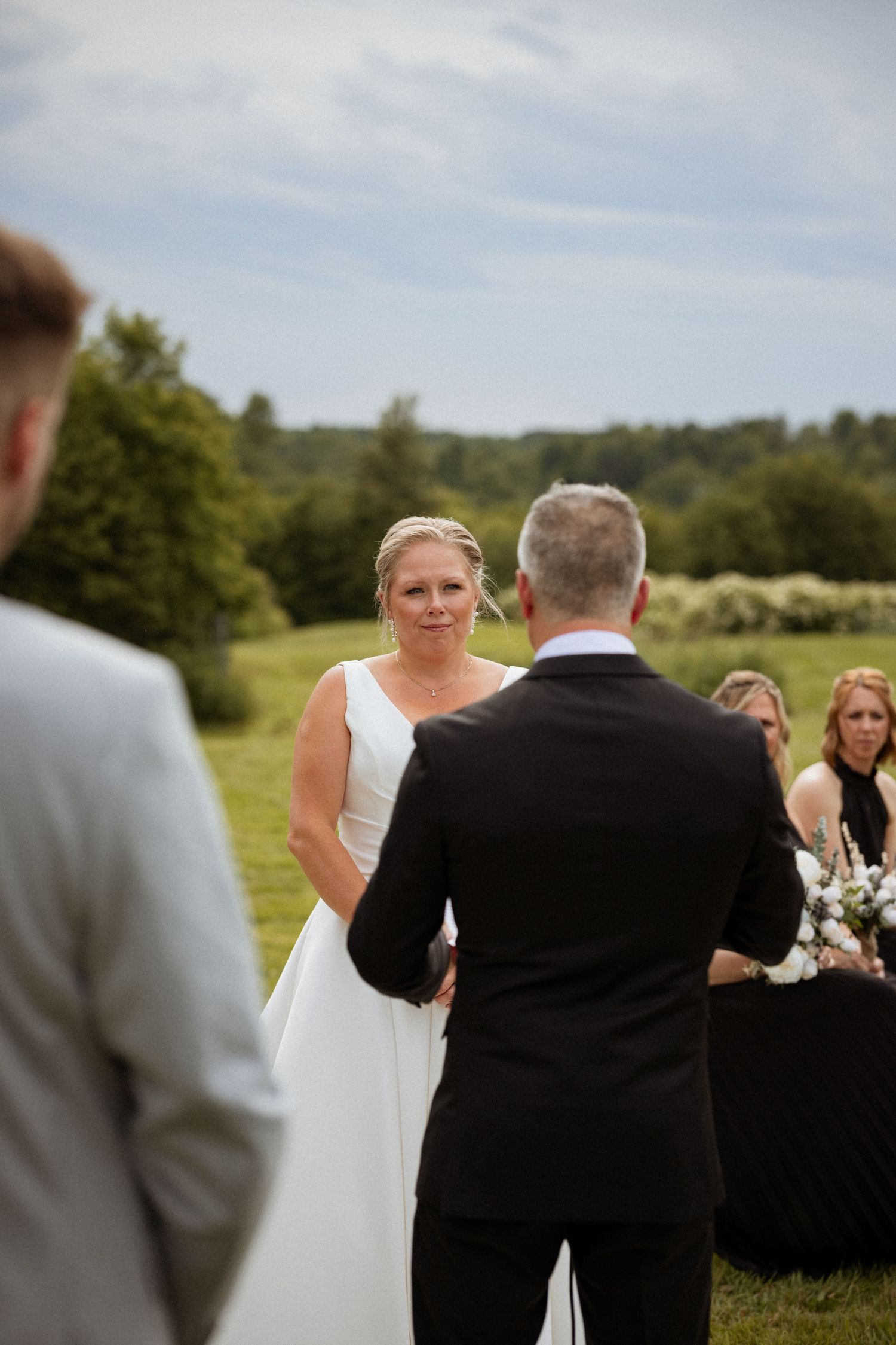 Bride in white gown exchanging vows with groom outdoors at a rustic wedding ceremony with green fields in the background.