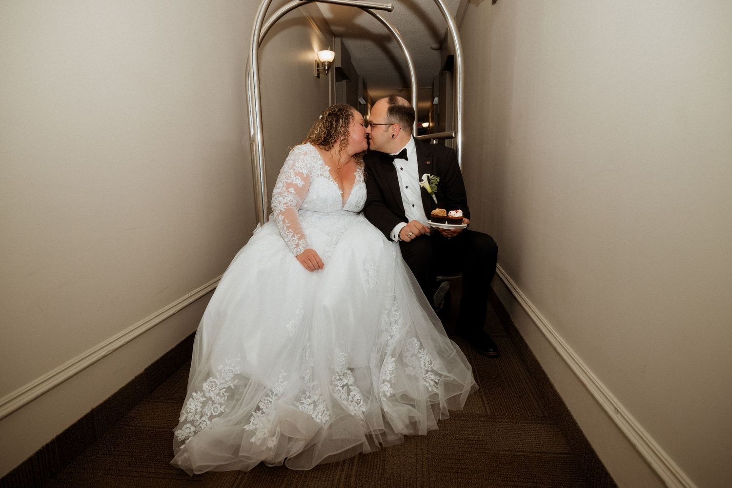 Bride in lace ball gown and groom in tuxedo share a tender kiss in an elegant arched hallway.