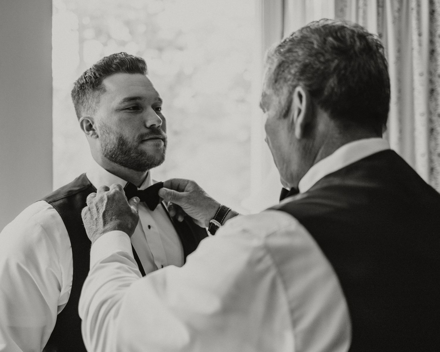 Black and white photo of groom having bow tie adjusted by older man before wedding ceremony.