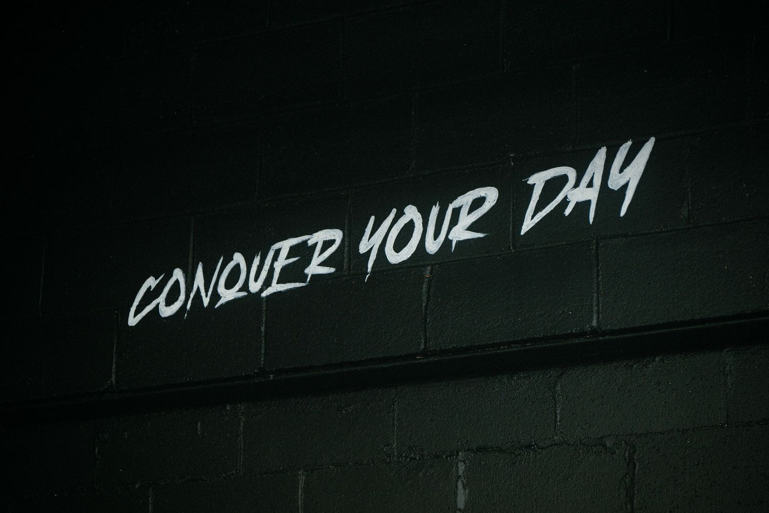 Motivational neon white graffiti text reading Conquer Your Day on dark brick wall.