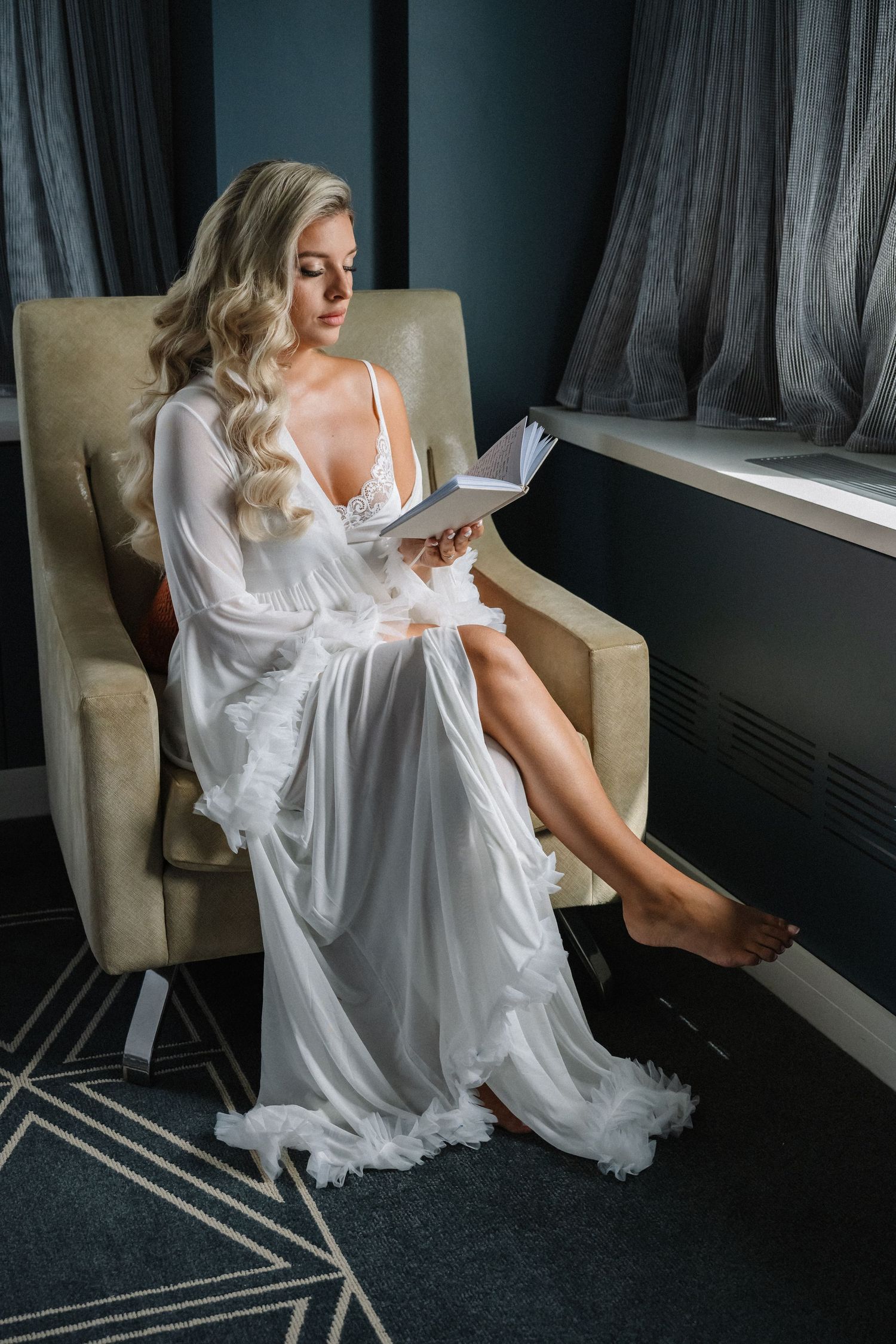 Elegant blonde woman in white lace bridal robe reading a book in an armchair by a window.