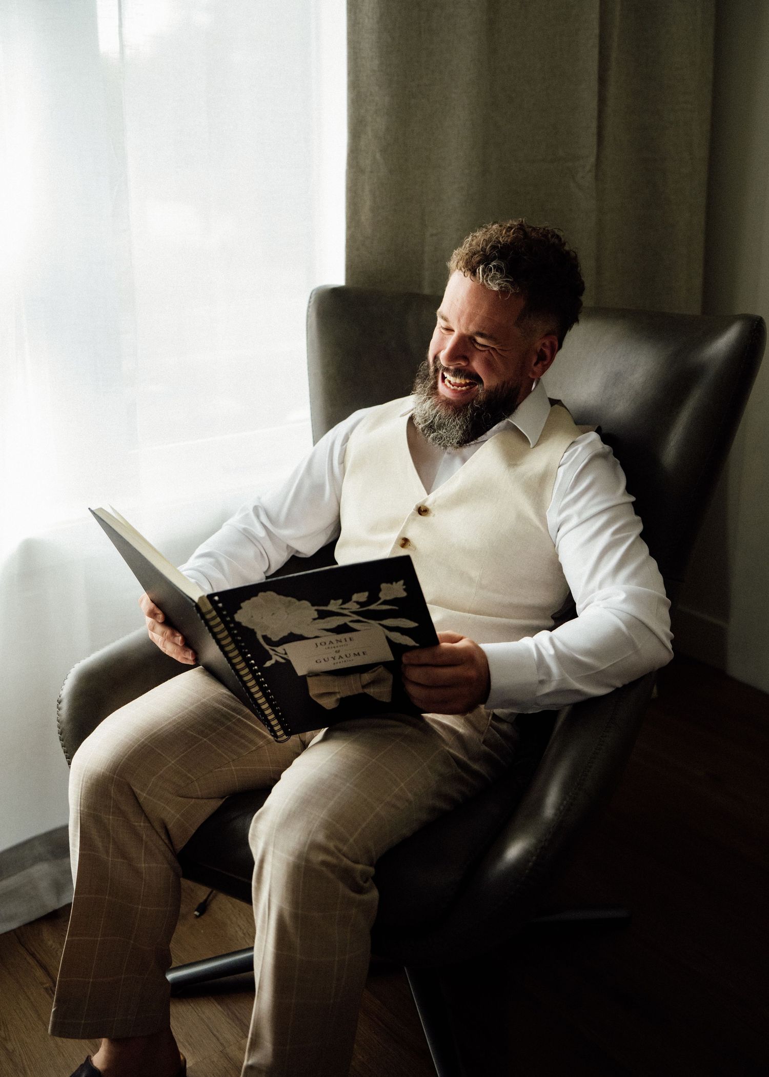 Happy bearded groom in white shirt and vest laughing while reading wedding album in chair.