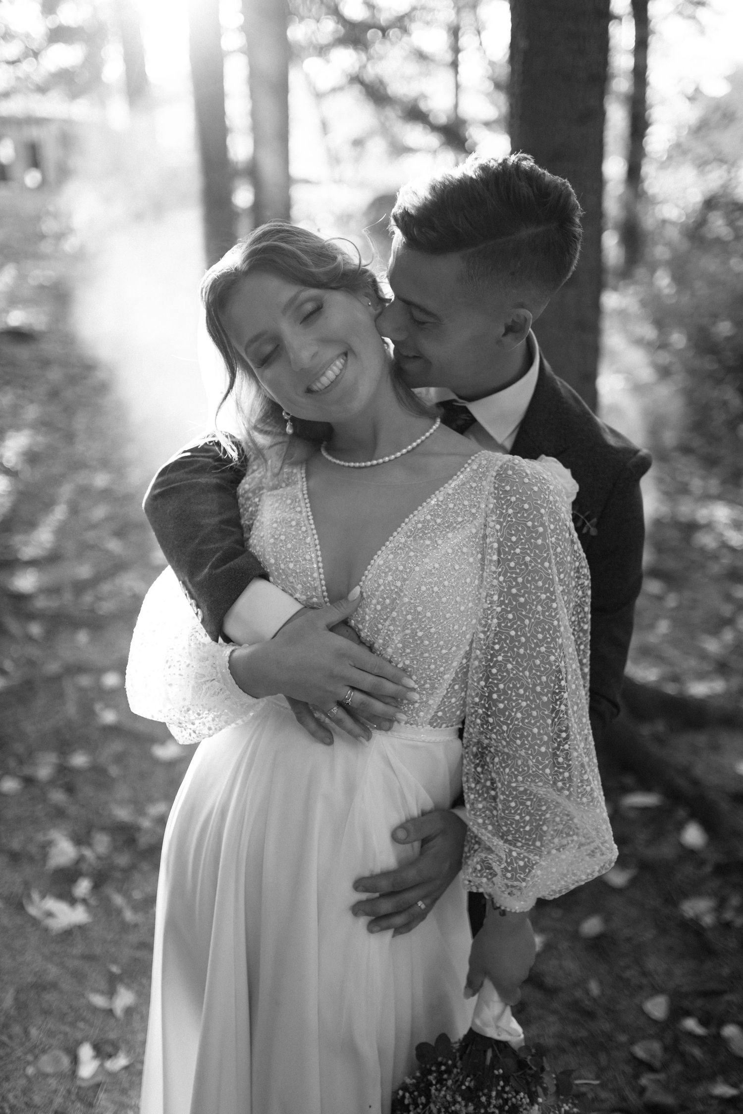 Black and white portrait of smiling bride in pearl gown embraced by groom in sunlit forest setting.