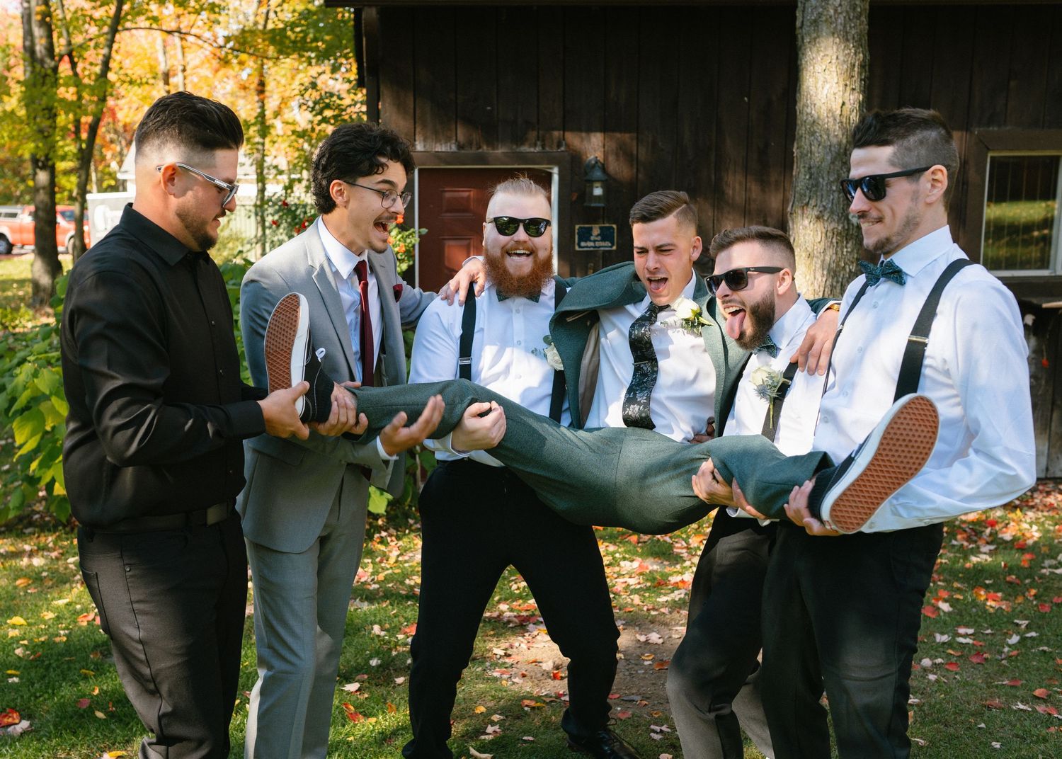 Groomsmen laughing and carrying groom outdoors at fall wedding near rustic barn.