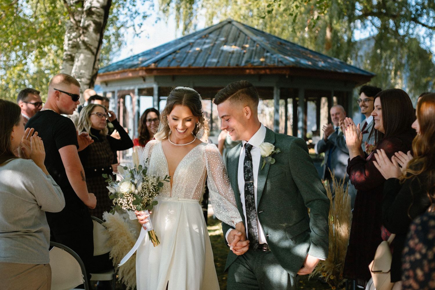 Newlyweds walk hand-in-hand through cheering guests after outdoor wedding ceremony.