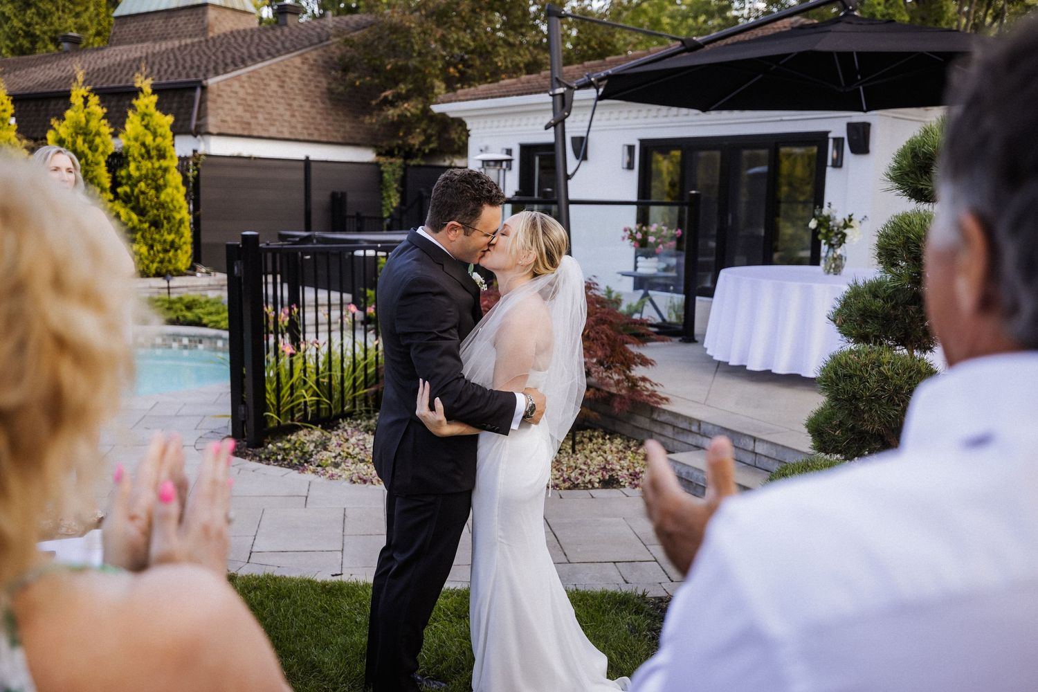 Newlyweds share first kiss as bride and groom while wedding guests applaud in backyard ceremony.